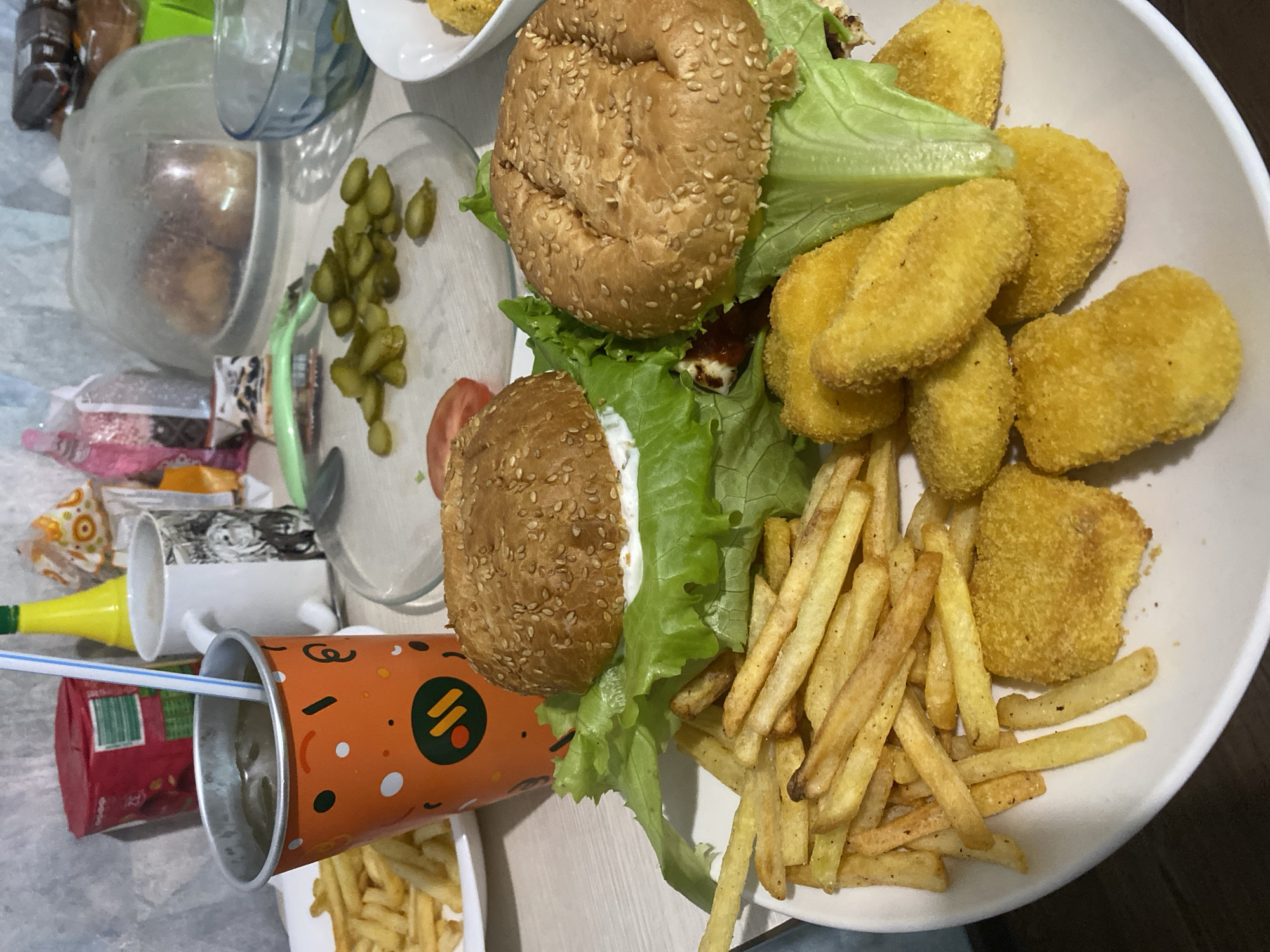 The image shows a table with various food items, suggesting a meal setting. Two burgers, with sesame seed buns, lettuce, and visible fillings, are present. A white plate contains fried chicken nuggets and french fries. Pickled cucumbers are visible on a plate. A partially visible orange paper cup with a straw lies on its side, and another mug is visible in the background, next to other items. The location information "Izhevsk, Russia" implies the image was taken in this city. No people are visible in the image.