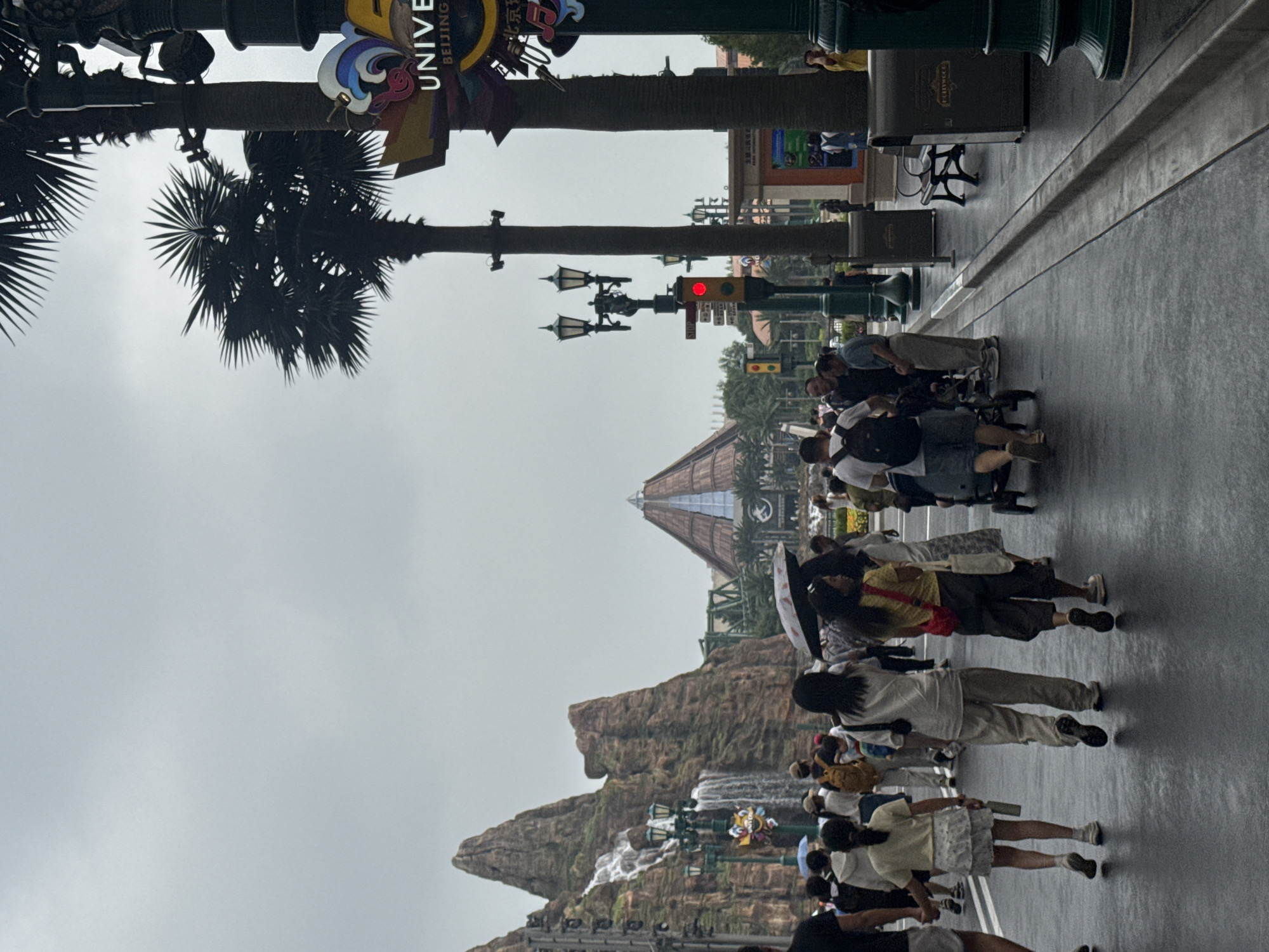 This image depicts a scene in Universal Studios Beijing, located in Tongzhou District, China. The sky is overcast, suggesting a cloudy or rainy day, as several people are carrying umbrellas. In the background, a large, pyramid-shaped structure with a thatched roof and glass panels is visible, along with a rocky mountain landscape featuring a waterfall. Palm trees are prominent in the foreground and middle ground. A streetlamp with two lanterns stands to the left of the frame, and a traffic light with a red signal is visible. A sign displaying "UNIVERSAL BEIJING RESORT" in English and Chinese characters is mounted on a lamppost. A group of people are walking on a wet asphalt pathway. Some are holding hands, while others are pushing strollers or carrying bags. The pathway is lined with planters and trash receptacles. The overall impression is of a busy theme park environment on a damp day.