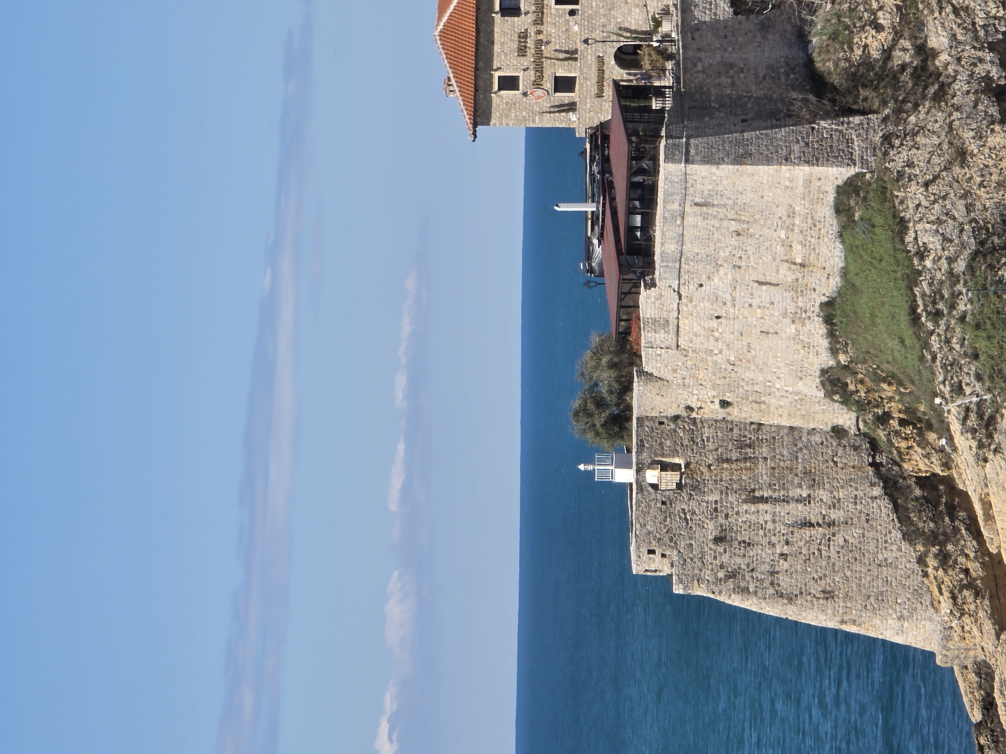The image depicts a vertical shot of a coastal scene in Ulcinj-Ulqin, Montenegro. The sky, featuring a few light clouds, and the sea dominate the left half of the image. The right half shows a rocky cliff supporting stone structures, one of which is identified as "HOTEL HEDONIST & RESTAURANT." A white lighthouse is visible on the wall below the hotel. Green vegetation grows sporadically on the cliff face.