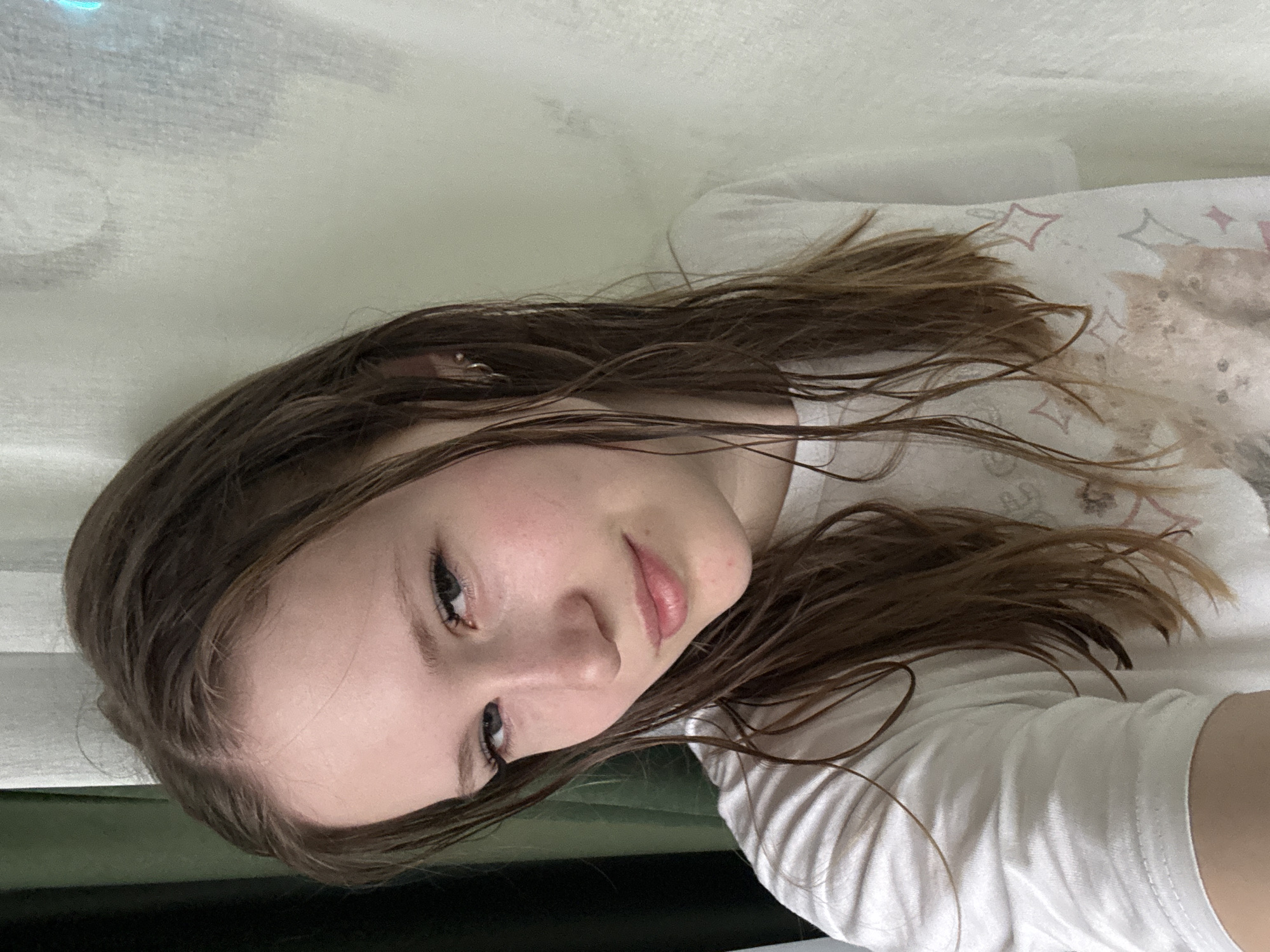 A young woman with light skin and brown hair is lying down, looking towards the camera. Her hair appears damp. She is wearing a white t-shirt with a faint pattern on the front. The background consists of a white surface. The location is not identifiable in this image.