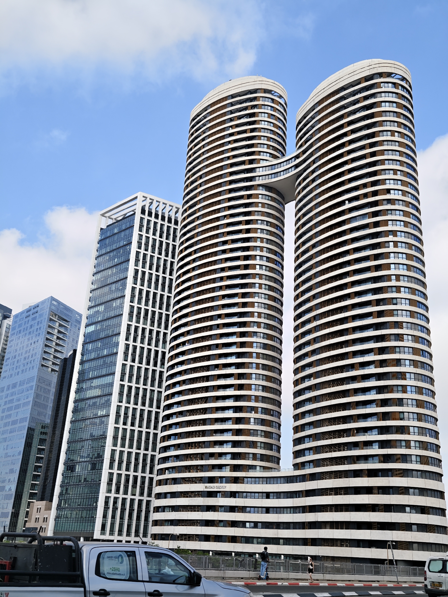 The image shows a daytime, exterior shot of the "Double Infinity" twin residential towers in Tel Aviv, Israel, as viewed from a street level. The towers are characterized by their unique curved architectural design, with horizontal bands of white and brown cladding interspersed with numerous windows. A sky bridge connects the two towers at a mid-level. In the foreground, a silver pickup truck is partially visible, with a sticker on its passenger side window. Further down the street, two individuals are visible on a walkway, one riding an electric scooter and the other walking, with a white van parked to the right. The sky is partly cloudy, with blue patches visible.