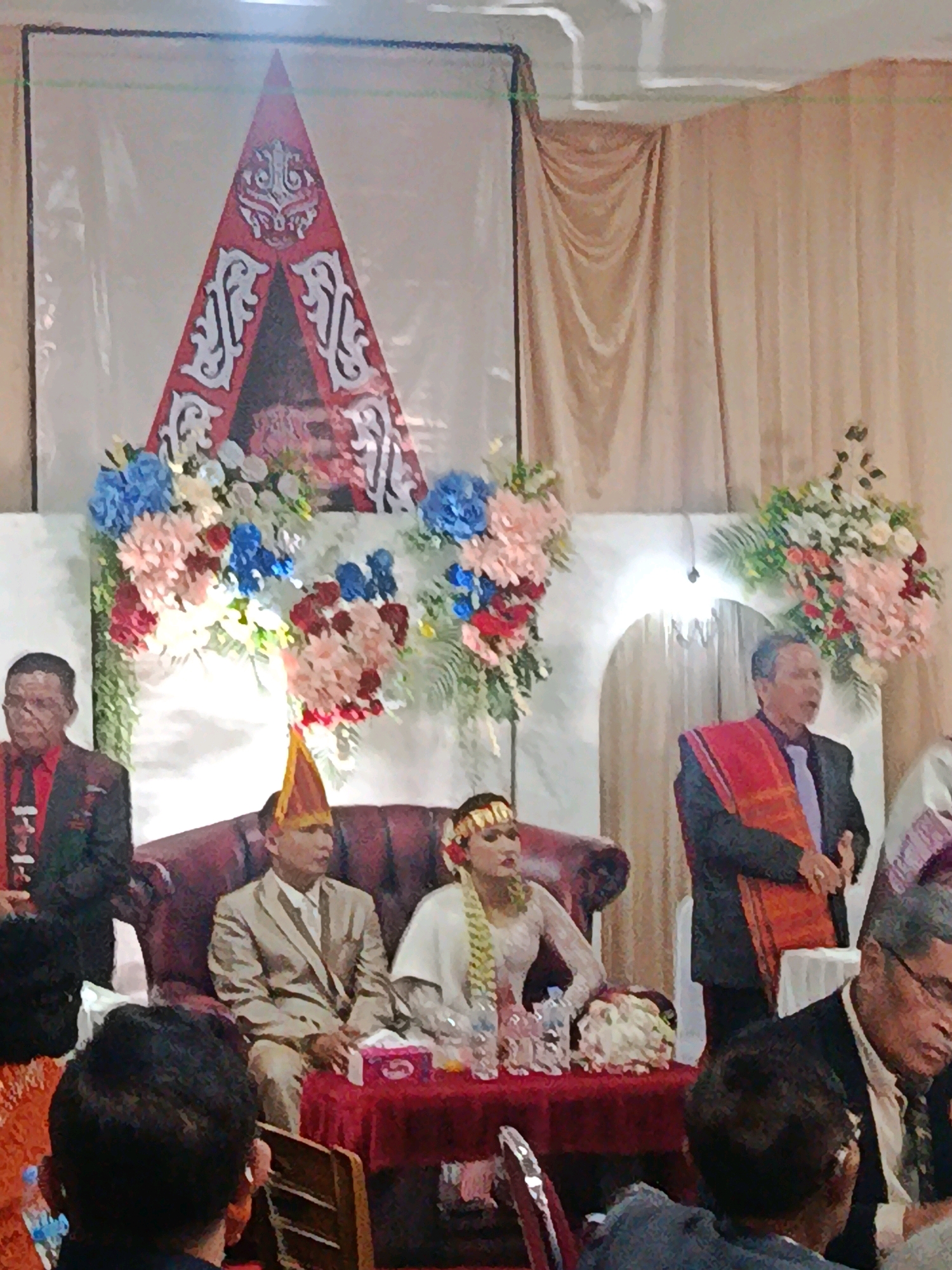 An indoor wedding ceremony is depicted in Babelan, Indonesia. A bride and groom are seated on a plush red sofa. The groom wears a cream-colored suit and a traditional conical hat. The bride is in a white wedding dress and a gold headpiece. To the left of the couple, a man in a black suit and red tie stands. To the right of the couple, another man in a black suit and a red sash over his shoulder stands and addresses the attendees. Behind the wedding party, a large red and white triangular backdrop with intricate designs is visible. The room is decorated with floral arrangements of pink, blue, and white flowers. A red tablecloth covers a table in front of the sofa, laden with wedding favors and glasses. The background features draped cream-colored curtains and a white archway. Several guests are partially visible in the foreground and to the right, observing the ceremony.