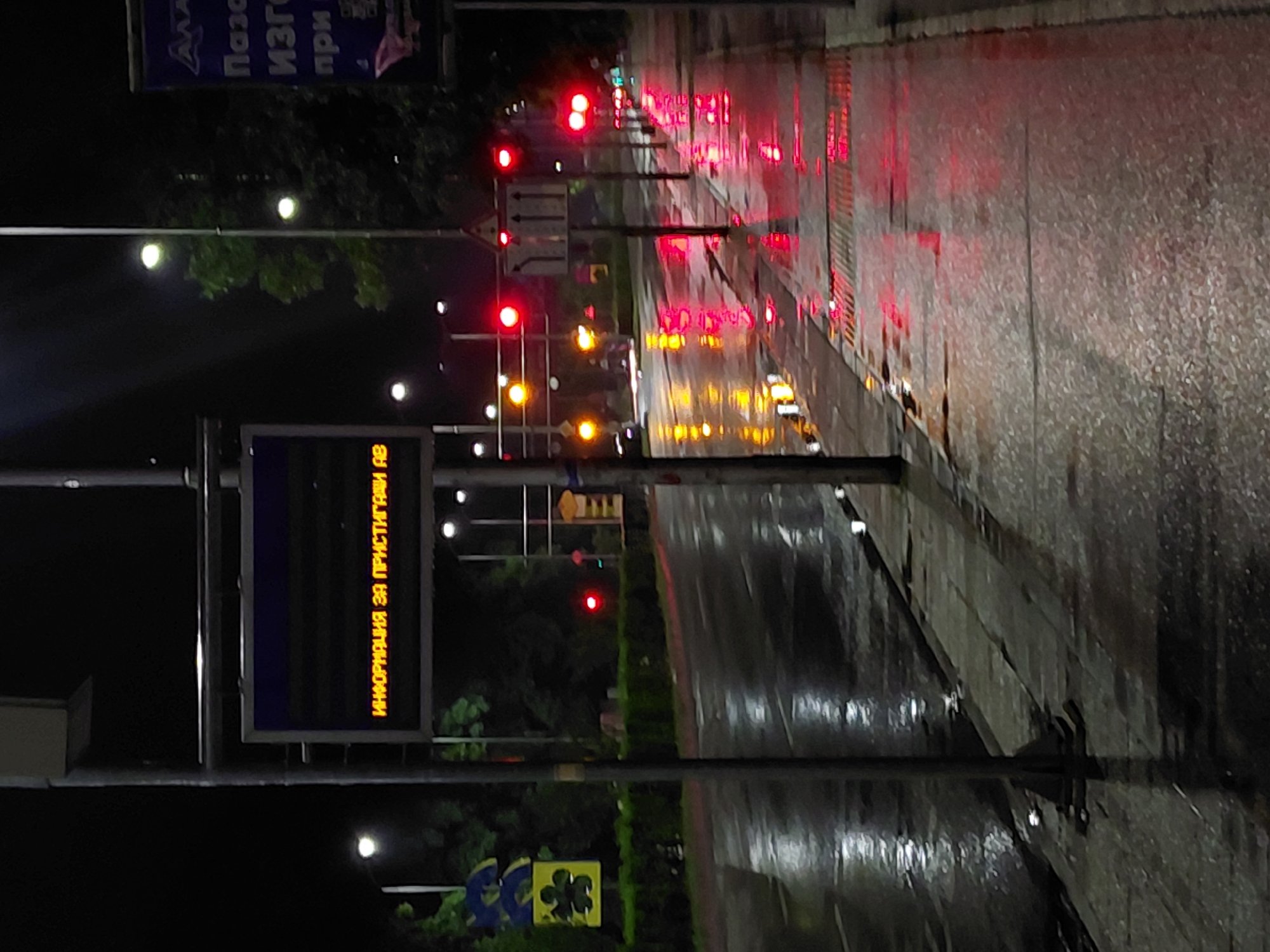 The image displays an urban street scene at night in the city of Unknown, Unknown, characterized by wet surfaces reflecting artificial light. The ground, encompassing both an asphalt road and a concrete sidewalk, is saturated with moisture, indicative of recent rainfall.
A central metal pole supports an illuminated electronic display board. The board exhibits orange Cyrillic text: "ИНФОРМАЦИЯ ЗА ПРИСТИГАЩИ АВ," which translates to "INFORMATION FOR ARRIVING VEHICLES/BUSES."
To the left of the central pole, a streetlight emits a bright orb of light, and a blue and yellow sign with an abstract, four-petal flower design is visible, partially obscured by dark foliage. The wet road surface beneath reflects these light sources.
To the right of the central pole, a series of operational red traffic lights extends into the distance along the street, casting vivid, elongated red reflections onto the wet sidewalk and road. A white, triangular street sign depicting three upward-pointing arrows is positioned centrally on the road. Further right, a large blue billboard with partially visible Cyrillic text is present. Multiple streetlights are also visible in the background, their light reflecting on the wet ground.
The scene is devoid of any individuals or actively moving vehicles.