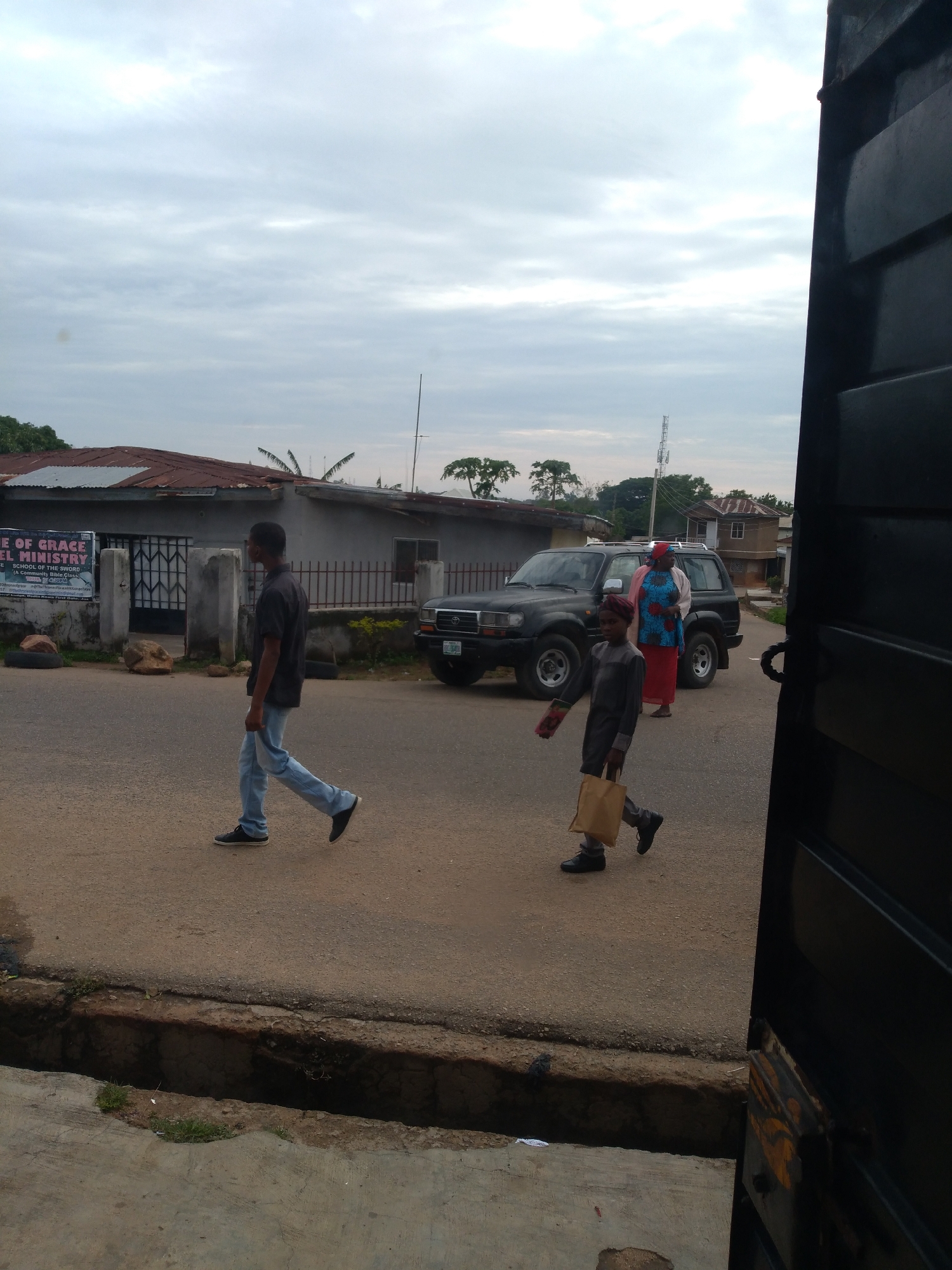 The image depicts a street scene in an urban or semi-urban environment under an overcast sky. The location is an unknown city.
In the foreground, a young child wearing a dark grey long-sleeved tunic, a dark red cap, and dark shoes walks towards the right side of the frame, carrying a light brown paper bag in their left hand. Behind the child, slightly to the left, a man wearing a dark short-sleeved collared shirt, light blue jeans, and dark shoes walks away from the viewer towards the left.
To the right, a dark-colored SUV, possibly a Toyota Land Cruiser, is parked on the roadside, facing left. Behind the parked SUV, a woman wearing a blue and red patterned top, a red skirt, and a light-colored head covering stands facing the left.
On the left side of the road, there is a single-story building with a rusty corrugated metal roof and grey walls. A white sign with blue text is affixed to its front, clearly displaying "HOUSE OF GRACE GOSPEL MINISTRY" and below it "SCHOOL OF THE SWORD". In front of this building, a low metal fence and large rocks are visible.
In the mid-ground and background, additional buildings with reddish roofs are scattered among green trees. A tall, thin cell tower or communication mast rises prominently in the background on the right side. The ground consists of a paved asphalt road, with an open concrete drainage ditch running parallel to the road in the immediate foreground.
On the far right of the frame, a large, dark black metal gate or door, possibly part of a building entrance, partially obscures the view.