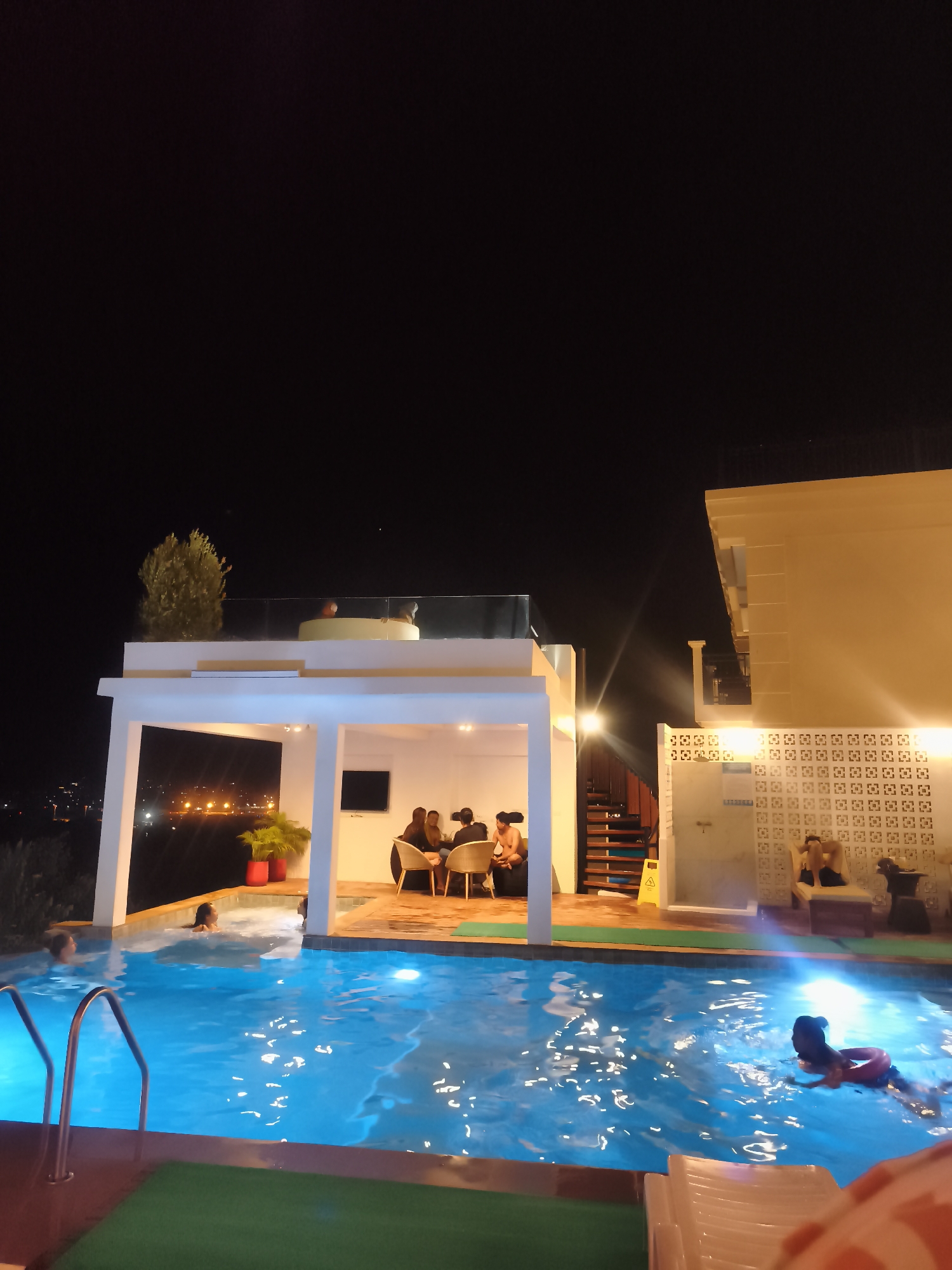 The image depicts an outdoor pool area at night in บ้านบางรักษ์, Thailand. The pool is illuminated with blue lights and has people swimming in it. To the left of the pool, there is a covered patio with a table where people are seated. A set of stairs lead to a rooftop area with a small tree and people relaxing. Adjacent to the patio, there's a building with decorative ventilation blocks and a sign labeled "TOILET", where people are sitting and relaxing. The overall scene suggests a relaxed, leisure setting at night.