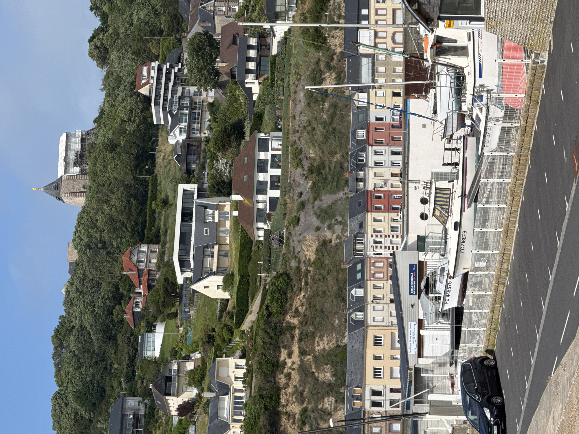 The image captures a panoramic view of Fécamp, France, showcasing a vibrant coastal town under a clear, sunny sky. The scene is divided between a lush, green hillside on the left and a more developed waterfront area on the right.
The hillside is densely covered with trees, interrupted by several modern residential buildings with sleek designs, flat roofs, and expansive windows. Further up the slope, a traditional church spire, topped with a golden figure, stands out, and some older houses with distinct red-tiled roofs are nestled among the foliage. One building on the hillside is surrounded by scaffolding, suggesting ongoing renovation or construction.
The right side of the image features a bustling waterfront. A paved road with white markings occupies the foreground, where a black car is parked. Behind a white metal fence, several boats are visible, some appearing to be dry-docked. Two large white boats prominent in the foreground bear the registration numbers "FC 716623" and "FC 445575," with "FC 52249" partially visible on another. These registrations, along with visible signage, confirm the location.
A building along the waterfront prominently displays "ÉCOLE MUNICIPALE DE VOILE DE FECAMP" (Municipal Sailing School of Fécamp) and a sign for "Canoë Kayak Fécamp Hautes Falaises" (Canoe Kayak Fécamp High Cliffs), connecting the scene to local maritime and outdoor activities. The strong shadows and bright blue sky indicate a sunny midday or early afternoon. The overall impression is that of a picturesque and active French coastal town, characterized by its natural landscape meeting urban development.