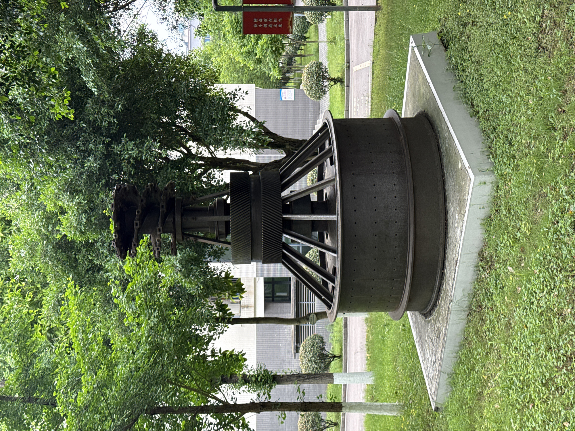 An eye-level outdoor shot shows a large, weathered, industrial-looking metal structure on a concrete platform surrounded by grass. The structure is cylindrical with a wide, flat rim and a grid of metal beams connecting the rim to a central hub with gears. To the left, trees obscure the structure and surrounding buildings. In the background, there is a pathway, more trees, and buildings. Based on available information, this is located in Jiangjin, China. A red sign is faintly visible in the top background, with Chinese characters on it.