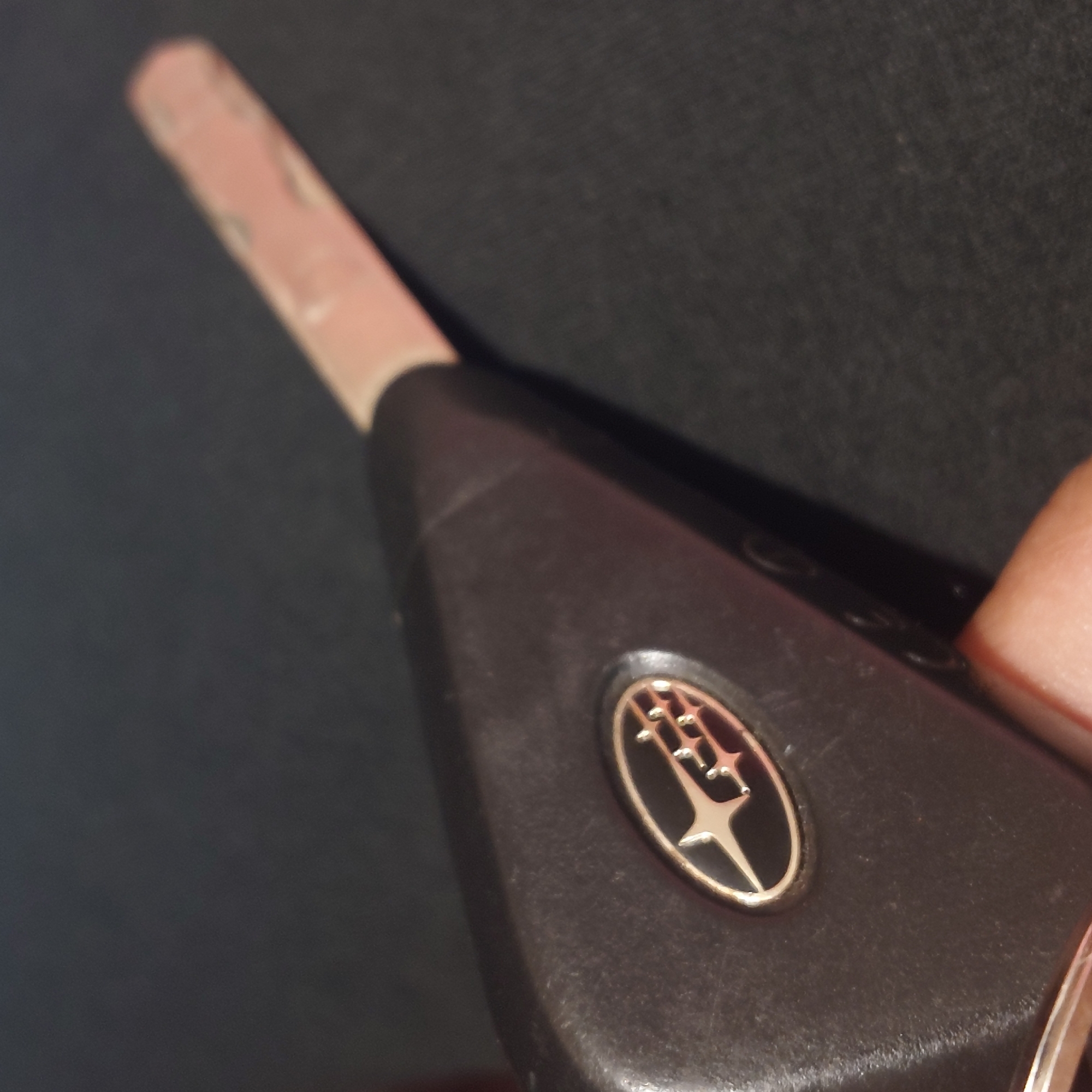 A close-up, angled shot shows a Subaru car key, black with a silver and gold emblem of the Subaru logo, lying on a dark, textured surface. The metal blade of the key, which appears to be a transponder key, is extended. The background is out of focus. There is no information about the location in the city of Belorechensk, Russia.