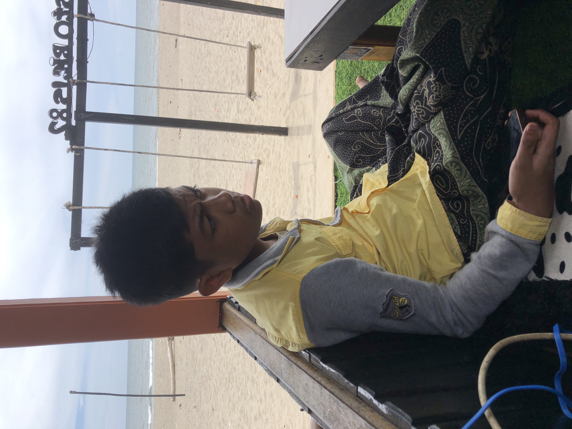 A young male with dark hair sits on a bench at a beachside cafe. He is wearing a yellow shirt with grey sleeves, and a patterned cloth draped over his legs. The beach has sand and the ocean is visible in the background, along with two swings hanging from a wooden frame. The cafe appears to be made of wood and has a roof. The overall scene suggests a relaxed atmosphere by the sea in Keudekruenggeukueh, Indonesia.