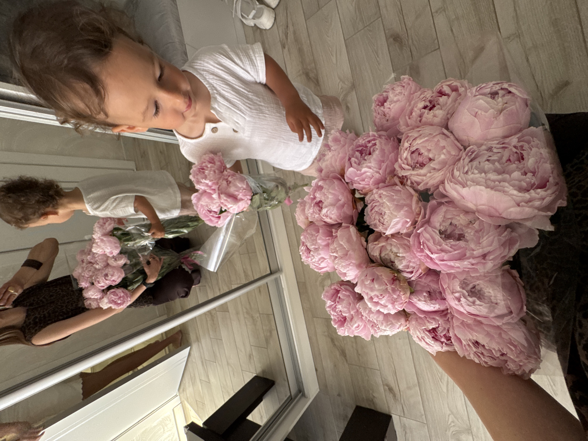 A young child with curly brown hair is standing in front of a mirror, holding a bouquet of pink peonies. The child is wearing a white button-down shirt and pink shorts. In the reflection, an adult is seen kneeling and holding another bouquet of pink peonies. The adult is wearing a leopard-print sleeveless top and dark pants. The room appears to be a home with light-colored wooden flooring and a large mirror. The context of Sochi, Russia is not present in the image.