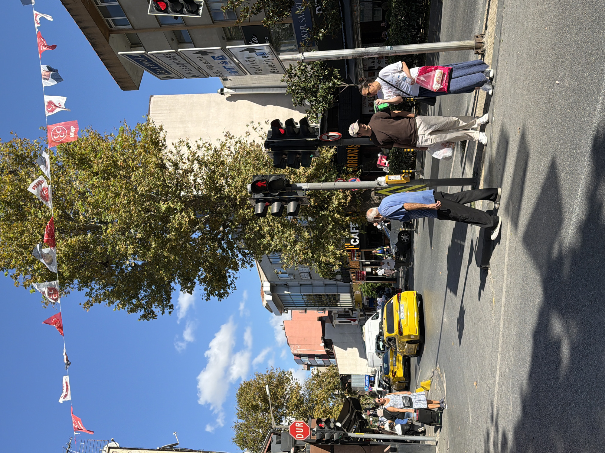 In Kadıköy, Turkey, a street scene unfolds under a bright, cloudy sky. A busy intersection features multiple yellow taxis, one with its license plate clearly visible (34 TCZ 263). Pedestrians navigate the crosswalk and sidewalks. A woman in a patterned dress walks with a rolling suitcase, and another woman in a white t-shirt and blue skirt stands with shopping bags. A man in a blue shirt and dark pants is crossing the street. In the background, buildings line the street, with signs indicating directions and businesses like "Cafe" and "GÖREKCİS". Overhead, a string of small flags or banners decorates the street, some displaying political party logos. Traffic lights control the flow of vehicles, with one showing a red signal for pedestrians and vehicles. A large tree with autumn foliage stands prominently near the center of the scene, casting shadows on the asphalt.