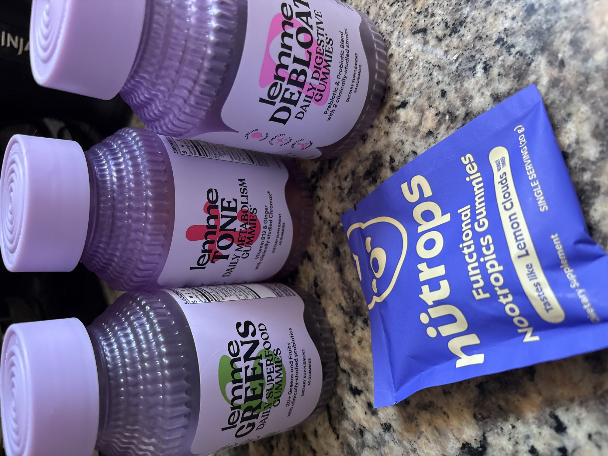 The image shows three bottles of "Lemme" brand gummy supplements, and a blue packet of "Nütrops" functional nootropics gummies, arranged on a speckled countertop. The Lemme bottles, each with a light purple lid, are partially visible. The top bottle appears to be "Lemme Debloat Daily Digestive Gummies." The middle bottle is "Lemme Tone Daily Metabolism Gummies." The bottom bottle is "Lemme Greens Daily Superfood Gummies." The Nütrops packet is labeled as "Functional Nootropics Gummies," lemon clouds flavor. The location is not discernible from the image.