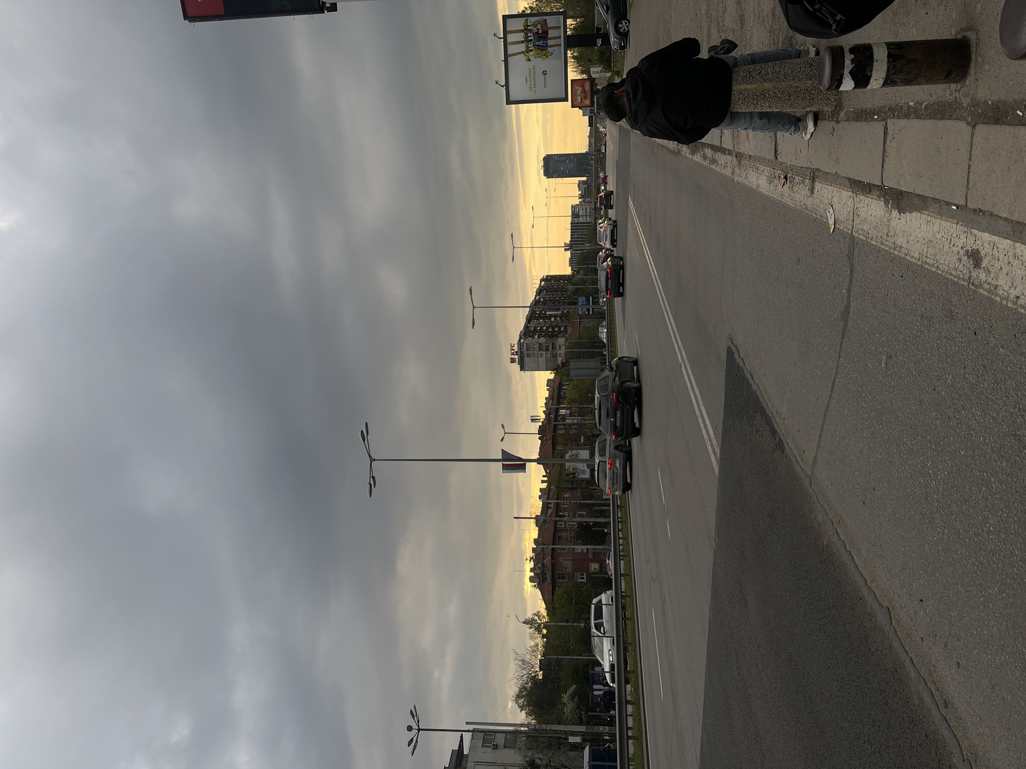 An urban street scene unfolds during what appears to be late afternoon or early evening, likely sunset, in Sofia, Bulgaria. The image captures a multi-lane asphalt road with white lane markings, populated by several cars, suggesting either moving traffic or a temporary stop.
On the right side of the frame, a person is partially visible, standing on a concrete sidewalk next to a low concrete barrier or post marked with black and white stripes. The individual wears a dark jacket and blue jeans. The road extends into the distance, lined by various buildings, some appearing residential and others commercial. Multiple streetlights with distinctive multi-arm designs are spaced along the thoroughfare.
The sky is largely covered with gray, overcast clouds, but a vibrant golden-yellow light illuminates the horizon, indicating the setting sun and casting a warm glow over the distant cityscape. Several large billboards are prominently displayed above the buildings. One billboard clearly shows the text "KFC", while another features Cyrillic text, including the phrase "е с грижа" (with care/carefully). The overall impression is that of a typical city street transitioning from day to night.
