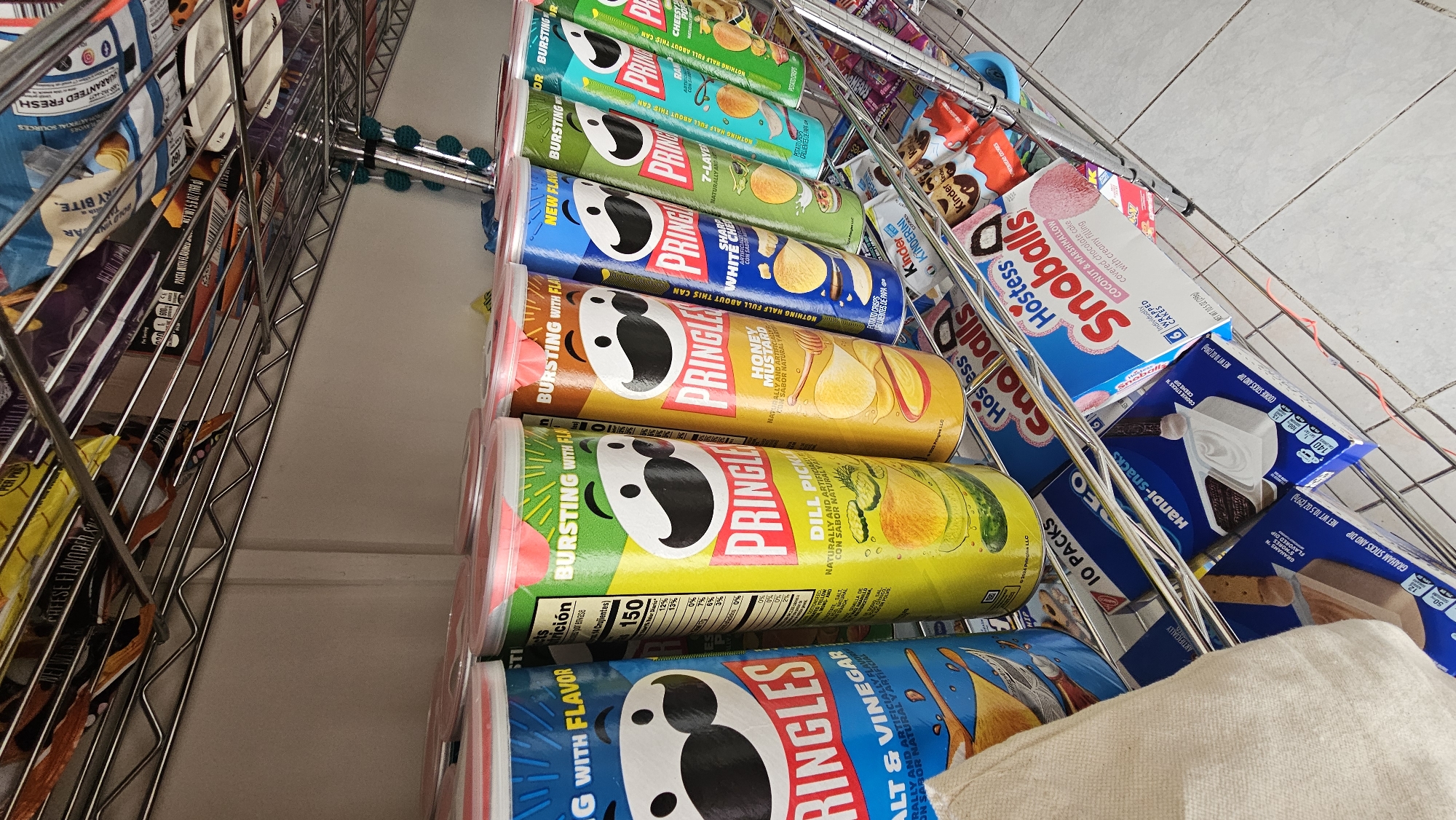 This is a close-up, angled shot of a metal shopping cart or shelving unit filled with various snack foods. Prominently featured are several cans of Pringles potato crisps in different flavors, including Dill Pickle, Honey Mustard, Sharp White Cheddar, 7-Layer Dip, and Salt & Vinegar. To the right of the Pringles are boxes of Hostess SnoBalls and Handi-Snacks. Additionally, there are packages of Kinder snacks visible. The lighting in the scene is bright and the image is taken at an angle, so it cannot determine the exact location of the city of San Bernardino Tlaxcalancingo, Mexico.