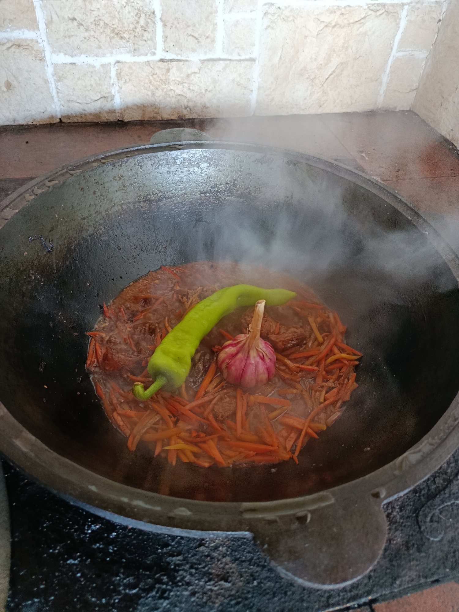 An overhead, close-up shot shows food being cooked in a large, dark, cast-iron cauldron. The cauldron is sitting on a dark, rough surface, possibly a stove or hearth. A light-colored brick wall is visible in the background. Inside the cauldron, a mixture of meat and julienned carrots is simmering in liquid, with a significant amount of steam rising from the dish. Resting on top of the food are a bright green chili pepper and a bulb of garlic. The scene appears to be outdoors or in an open-air kitchen area. There is no indication of the specific location within Misaylovo, Russia, other than it being a setting where traditional cooking methods might be employed.
