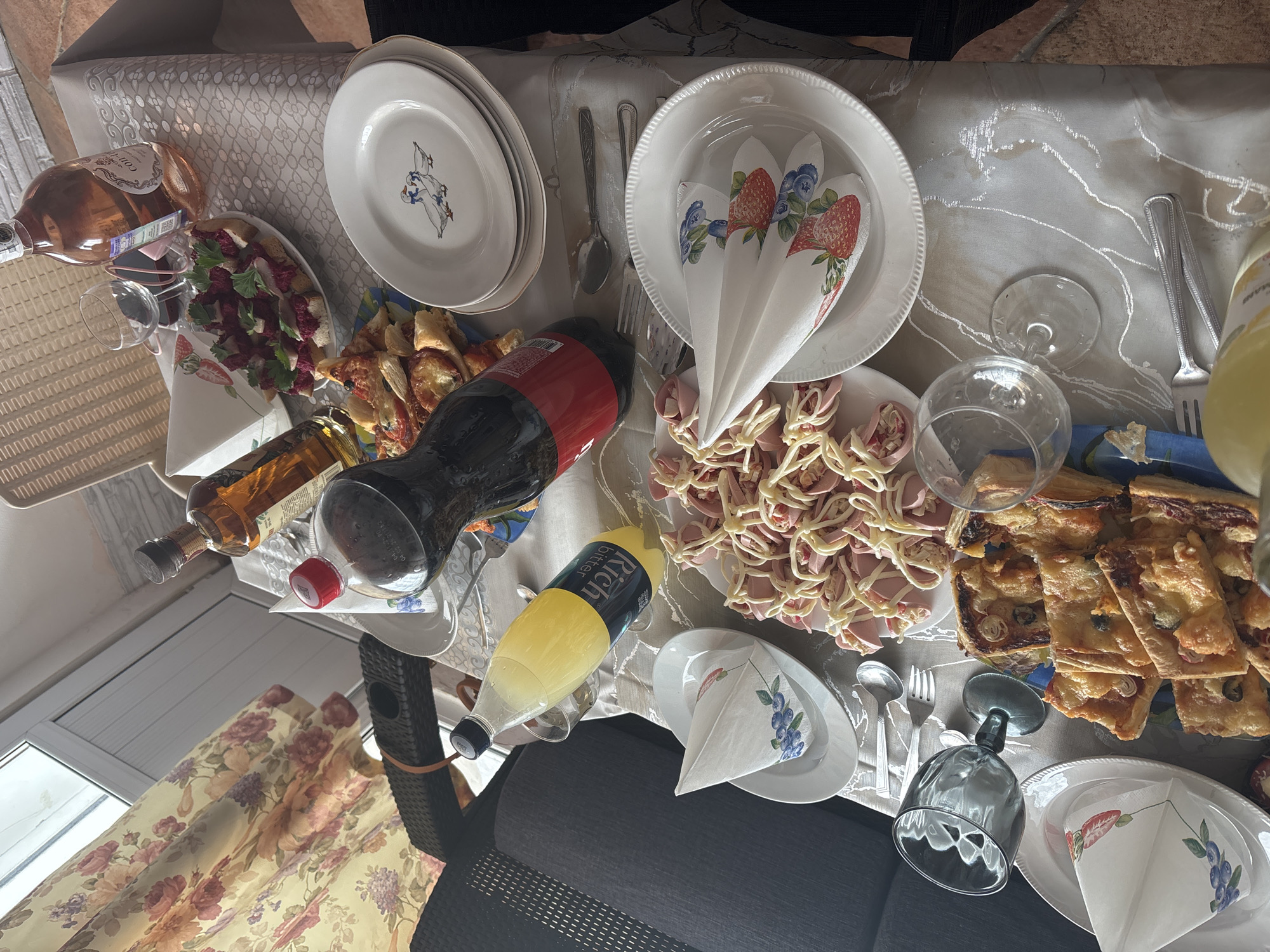 The image depicts a table laden with food and beverages, suggesting a gathering or meal. There are plates stacked and arranged with napkins, cutlery, and glasses. Bottles of liquids, including what appears to be Coca-Cola, a yellow-colored beverage in a bottle labeled "Rich Bitter," and a bottle of rosé wine, are visible. Food items include sliced ham with a cream-colored sauce drizzled on top, what appears to be pizza squares, and a salad. The tablecloth has a subtle pattern, and the overall scene suggests an indoor setting, possibly a dining room or kitchen. The presence of a window with a floral-patterned curtain in the background suggests a residential environment. The location is stated to be Sochi, Russia, though there is no clear indication within the image itself to confirm this.