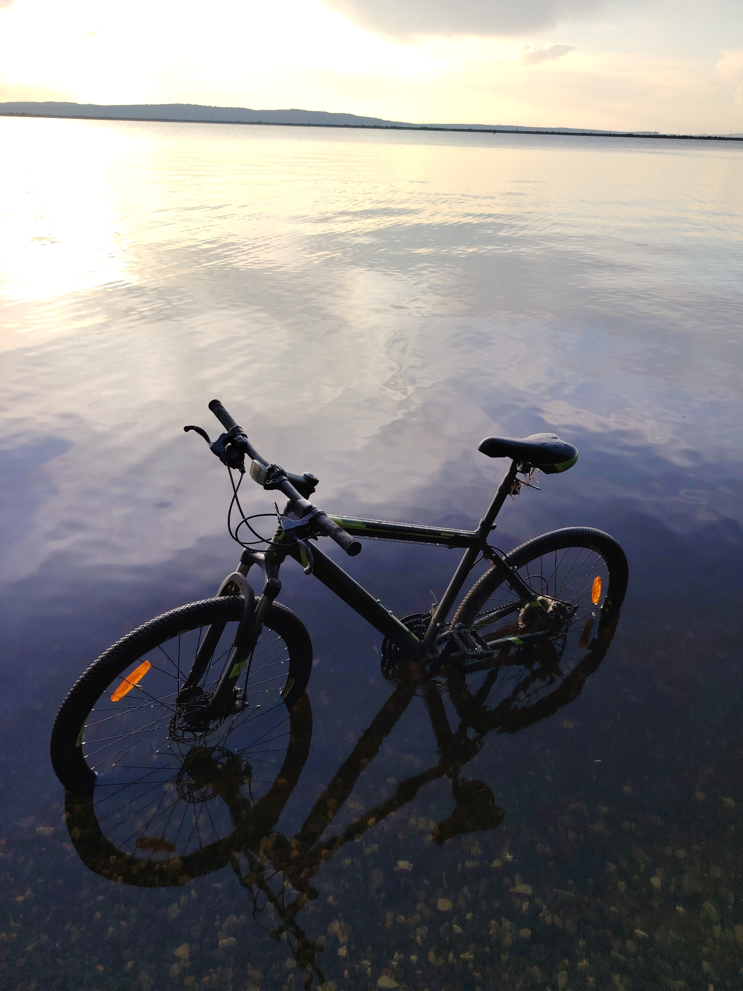 A dark-colored bicycle with green accents is partially submerged in shallow, calm water. The bicycle is positioned with its front wheel to the left and rear wheel to the right, angled slightly away from the viewer. The handlebars, seat, frame, wheels, and pedals are visible. The bicycle's reflection is clearly visible in the water, creating a distorted, darker image below. The water is shallow enough that the rocky, pebbly bottom is visible beneath the surface. In the background, a wide expanse of water stretches to the horizon, meeting a hazy sky with subtle cloud formations. The sky appears to be illuminated by either sunrise or sunset, casting a soft, warm glow on the water's surface, creating ripples and reflections of the sky. The overall scene is tranquil and evokes a sense of stillness. The location is identified as the Kama River in Chaikovsky, Russia.