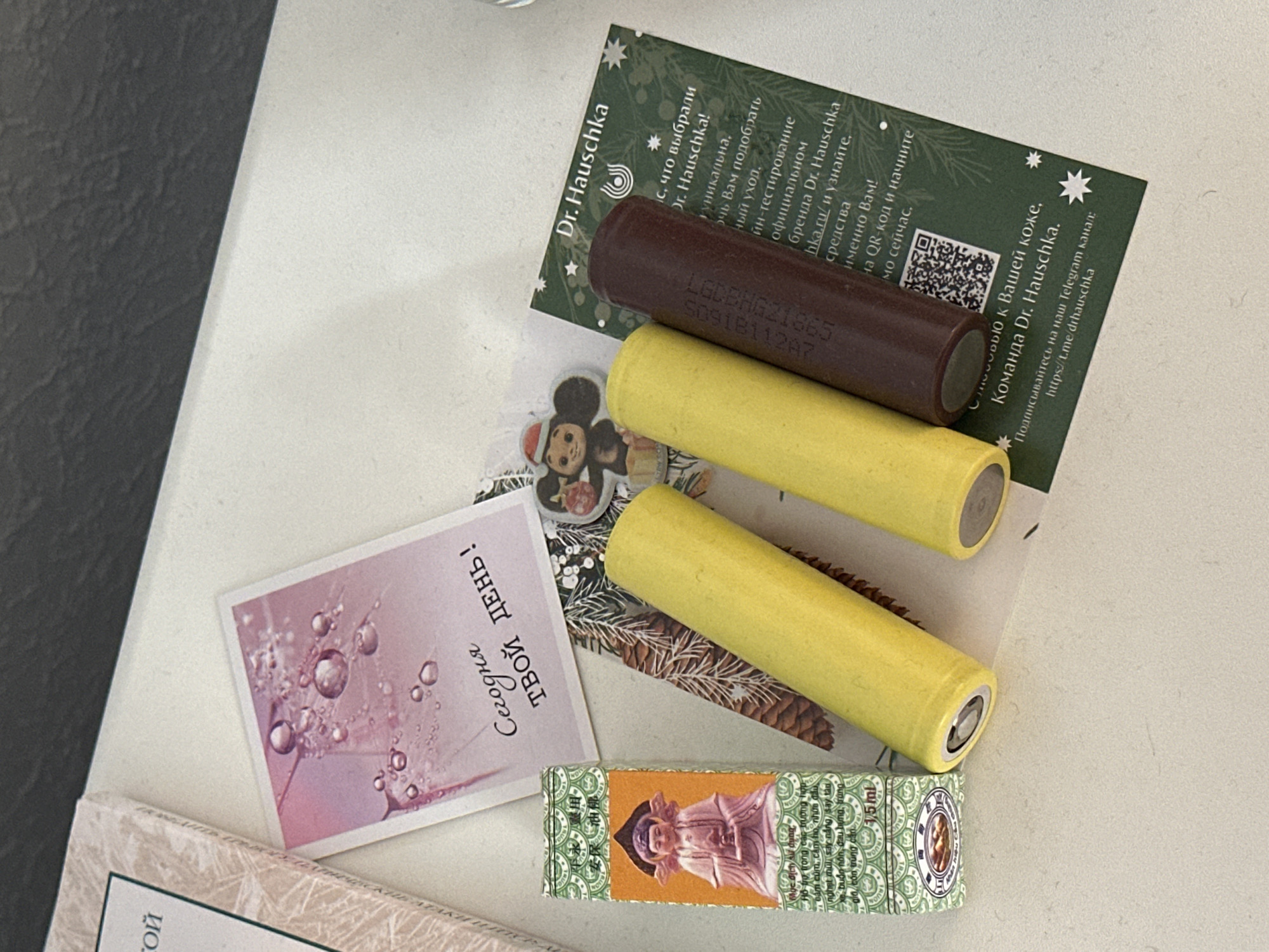 The image shows a collection of small objects on a white surface, possibly a desk or shelf. In the foreground, there are two cylindrical objects, one yellow and one dark brown, positioned diagonally. To the left of these cylinders is a small, rectangular green and orange box with text and an image of a seated figure. Behind these items, a pink card with droplets and text is partially visible, along with a small sticker of a monkey wearing a Santa hat. To the right, a green card with a star pattern and text, including "Dr. Hauschka" and a QR code, is present. The overall scene is a still life arrangement of various small items. The image does not provide any information about the city of Nevinnomyssk, Russia.