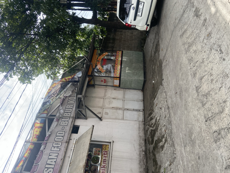 A street-level view of a small, white building in Baleendah, Indonesia, with a partially collapsed awning and a sign reading "Asian Food- Bubur Kompl". Next to the building is a smaller food stall with a sign that reads "Pondok Minang Masakan Padang". A white car with a license plate reading "D 1697 ZA" is parked in front of the stall. The scene is on a paved street in front of the building. The buildings are surrounded by overgrown vegetation. The image is taken from the street level looking toward the buildings. The sun is shining, and the sky is clear. The street is empty, with no people in sight.