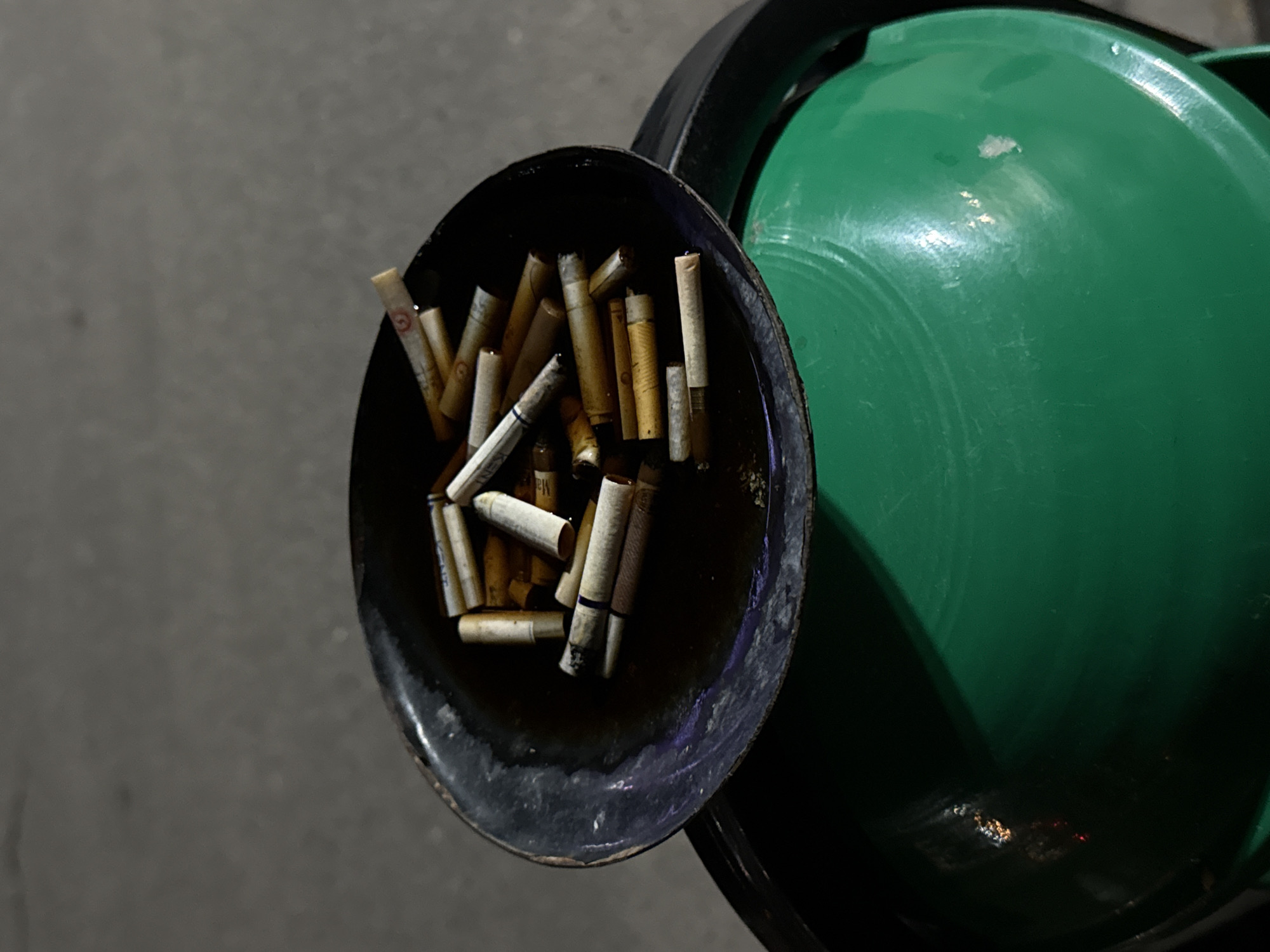 An overhead shot captures a black, circular ashtray overflowing with numerous cigarette butts, positioned to the left of a bright green, domed trash can lid. The ashtray, appearing to be made of metal, contains a dark liquid, likely water or residue, in which many cigarette filters are partially submerged. The filters are predominantly beige and white, with some showing faint markings, including a red circular logo with an indistinct letter (possibly 'G') on one, and faint blue bands or partial letters like 'EN' or 'PM' on others, none clearly legible to identify a brand.
The green trash can lid, visible on the right, is shiny and reflects some light, indicating a smooth, possibly plastic or painted metal surface. Its vibrant color contrasts with the darker tones of the ashtray and the subdued grey background, which appears to be a paved or concrete surface.
The lighting is dim, suggesting the image was taken at dusk, night, or in an indoors setting with low artificial light. No people are visible, nor is any specific activity taking place beyond the collection of cigarette discards. The scene depicts a common public smoking area setup, with the ashtray placed conveniently next to a bin for waste disposal. No definitive location cues are present within the image itself.