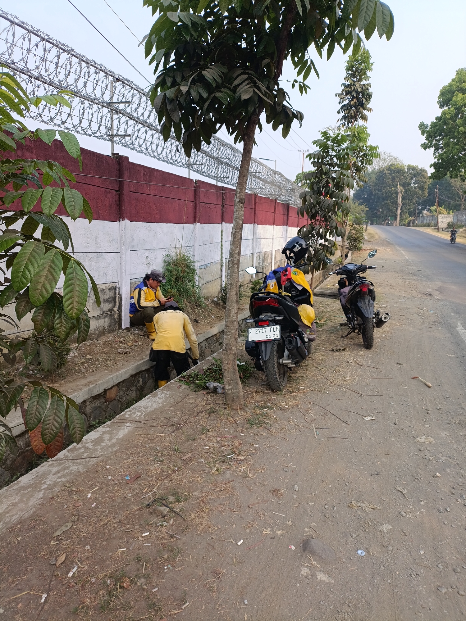 In Cariu, Indonesia, two individuals, presumably construction or maintenance workers, are observed near a roadside ditch. The taller individual, wearing a yellow shirt, black pants, and yellow boots, is actively working in the ditch. The second individual, in a blue and yellow jacket, olive green pants, and yellow boots, is seated on the edge of the ditch, looking at their phone. Both individuals wear hats. Two motorcycles are parked on the dirt shoulder of the paved road adjacent to the ditch. A red and white concrete wall topped with barbed wire fencing runs along the left side of the scene. Trees and foliage are present throughout the environment. The road extends into the background, with a distant figure on a motorcycle. The ground is a mix of dirt and debris, including cigarette butts. The sky is clear and bright.