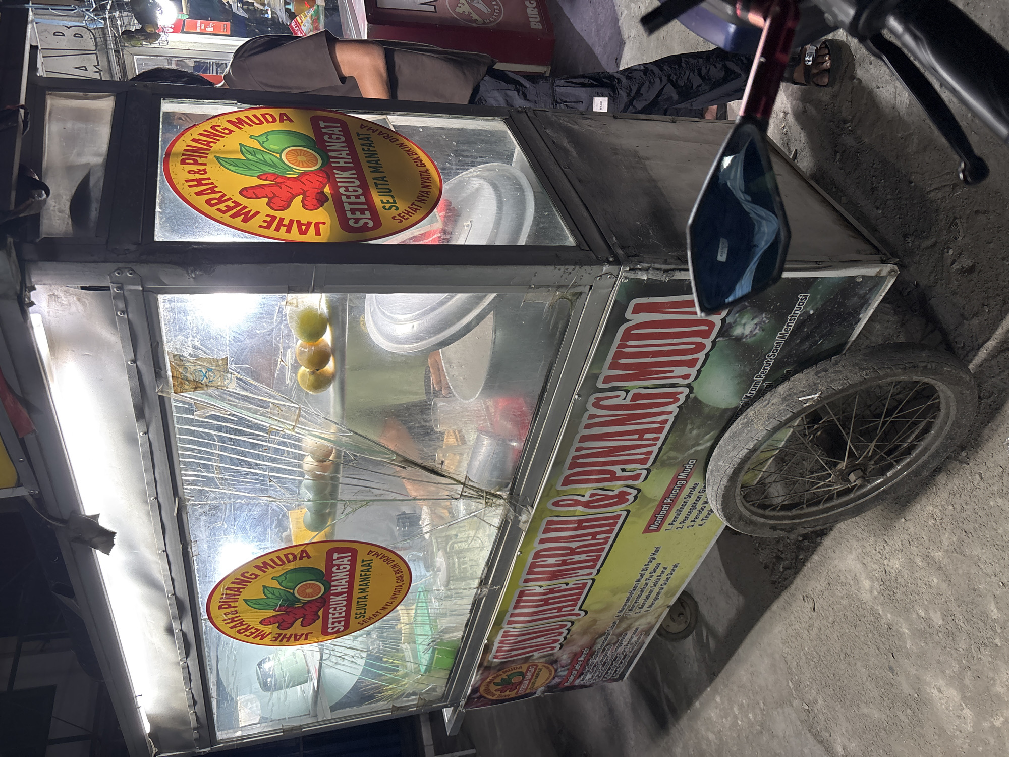 A street vendor's cart selling "Susu Jahe Merah & Pinang Muda" is parked on a dusty, unpaved surface in the city of Bekasi, Indonesia. The cart is illuminated by overhead lights and features a glass display case filled with ingredients and containers. Prominently displayed on the cart are circular signs with red and yellow graphics and text in Indonesian, advertising the drinks and their supposed health benefits. A large banner along the bottom of the cart also lists the benefits in Indonesian. A person, partially obscured, stands behind the cart, wearing dark pants and a brown shirt. The foreground shows a motorcycle's mirror and handlebars, suggesting the vendor or a customer arrived by motorcycle. The overall environment appears to be a roadside stall in a modest urban area.