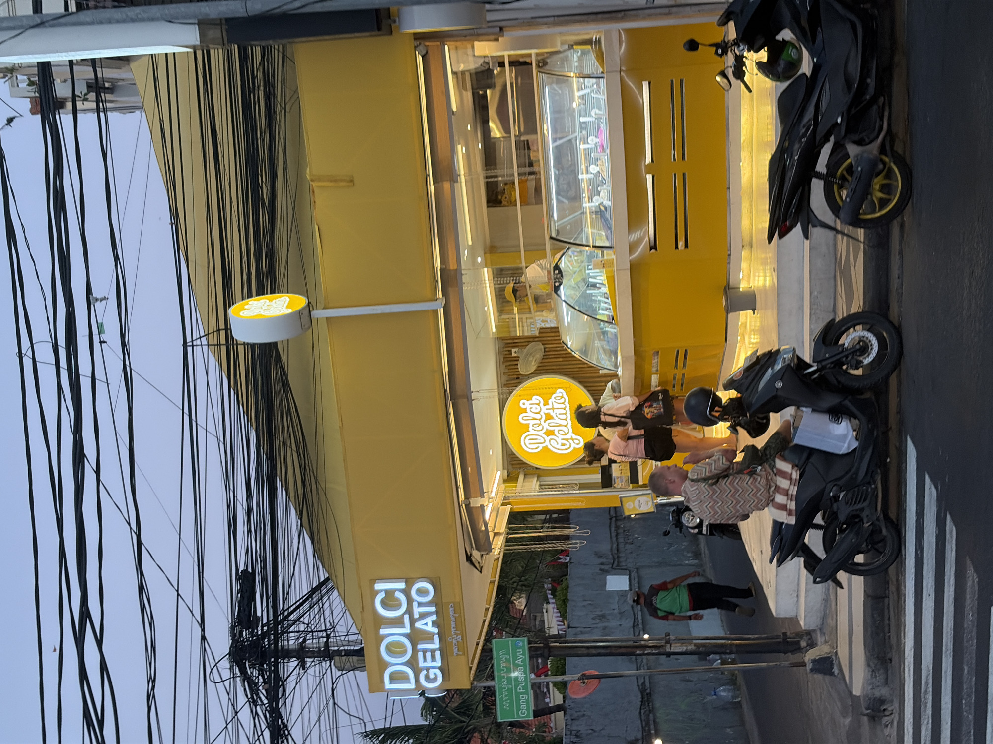 The image depicts the exterior of "Dolci Gelato," a bright yellow gelato shop located in Kuta, Indonesia. The shop features a large, illuminated sign with the name "DOLCI GELATO" in white block letters. Above the entrance, a circular neon sign also reads "Dolci Gelato" in a stylized font. The shop's facade is primarily yellow, with glass display cases showcasing gelato.
In the foreground, two motorcycles are parked on the street. A man wearing a patterned shirt sits on one of the motorcycles, facing away from the camera. Another motorcycle is parked to the right, and a green helmet rests on its seat.
Several people are visible near the entrance of the shop. Three women are standing at the counter, appearing to be ordering or receiving gelato. A man in a green and black shirt walks past the shop on the sidewalk, carrying what appears to be a plastic water bottle.
The street in front of the shop is paved, and a pedestrian crosswalk with white stripes is visible in the lower part of the image. A green street sign with white lettering indicates "Gang Puspa Ayu," identifying the specific street or alley. Overhead, a dense network of electrical wires is strung across the sky, a common sight in this region. The lighting suggests it is dusk or early evening, with artificial lights from the shop illuminating the scene. The overall atmosphere is that of a typical street scene in a bustling urban area of Kuta.