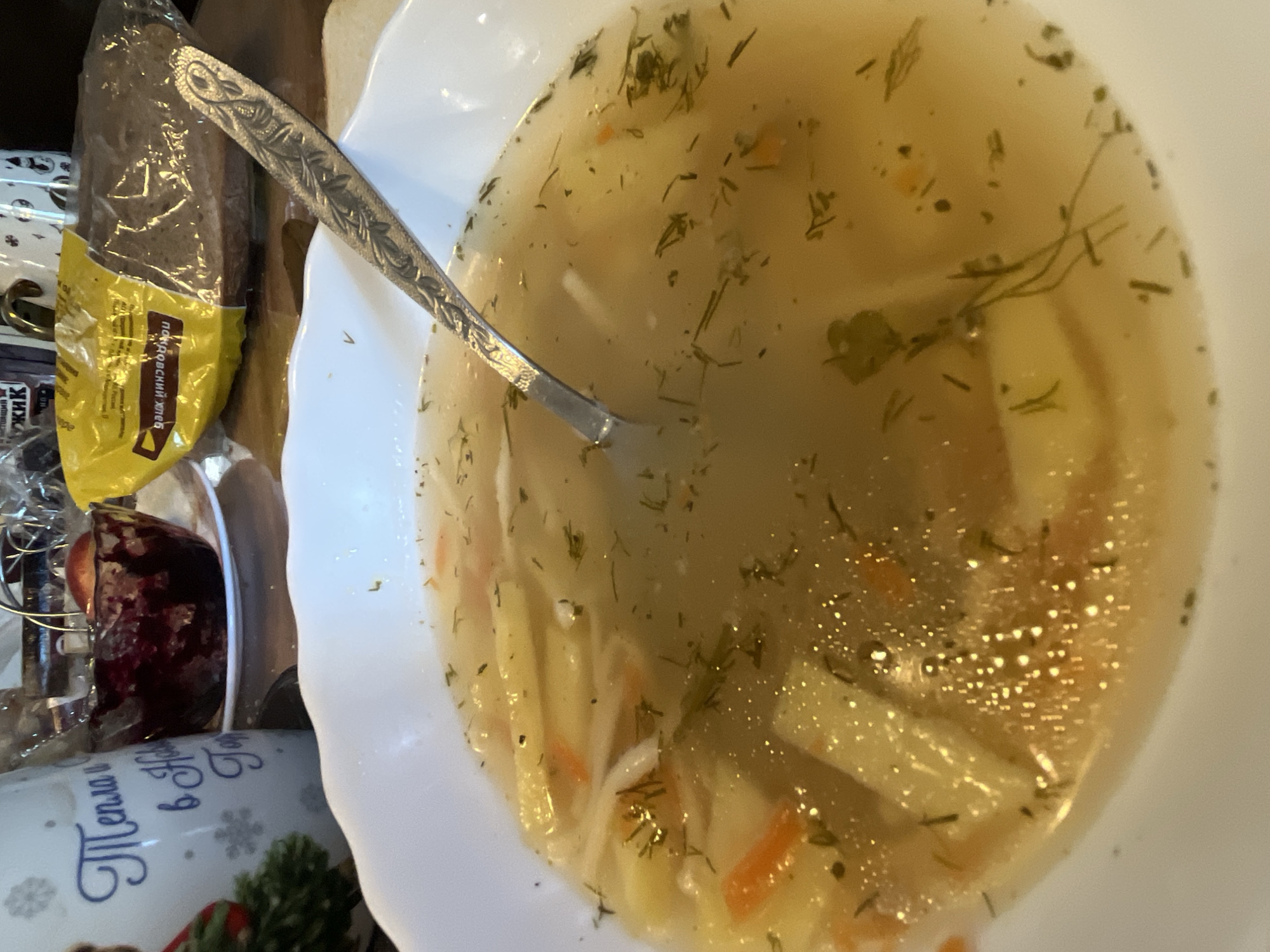 A white plate of soup with potatoes, carrots, noodles, and dill is on a table, a spoon is in the soup. A loaf of bread in a yellow package and a mug are next to the plate. The mug has a blue inscription in Russian: "Tepla v Novyy God". This scene is likely to be a meal setting. The location is not discernible from the image. The text on the packaging of the bread also contains Russian text, suggesting a region of origin, but not specifically Chaikovsky. Without further information, it's impossible to pinpoint the location to Chaikovsky, Russia.