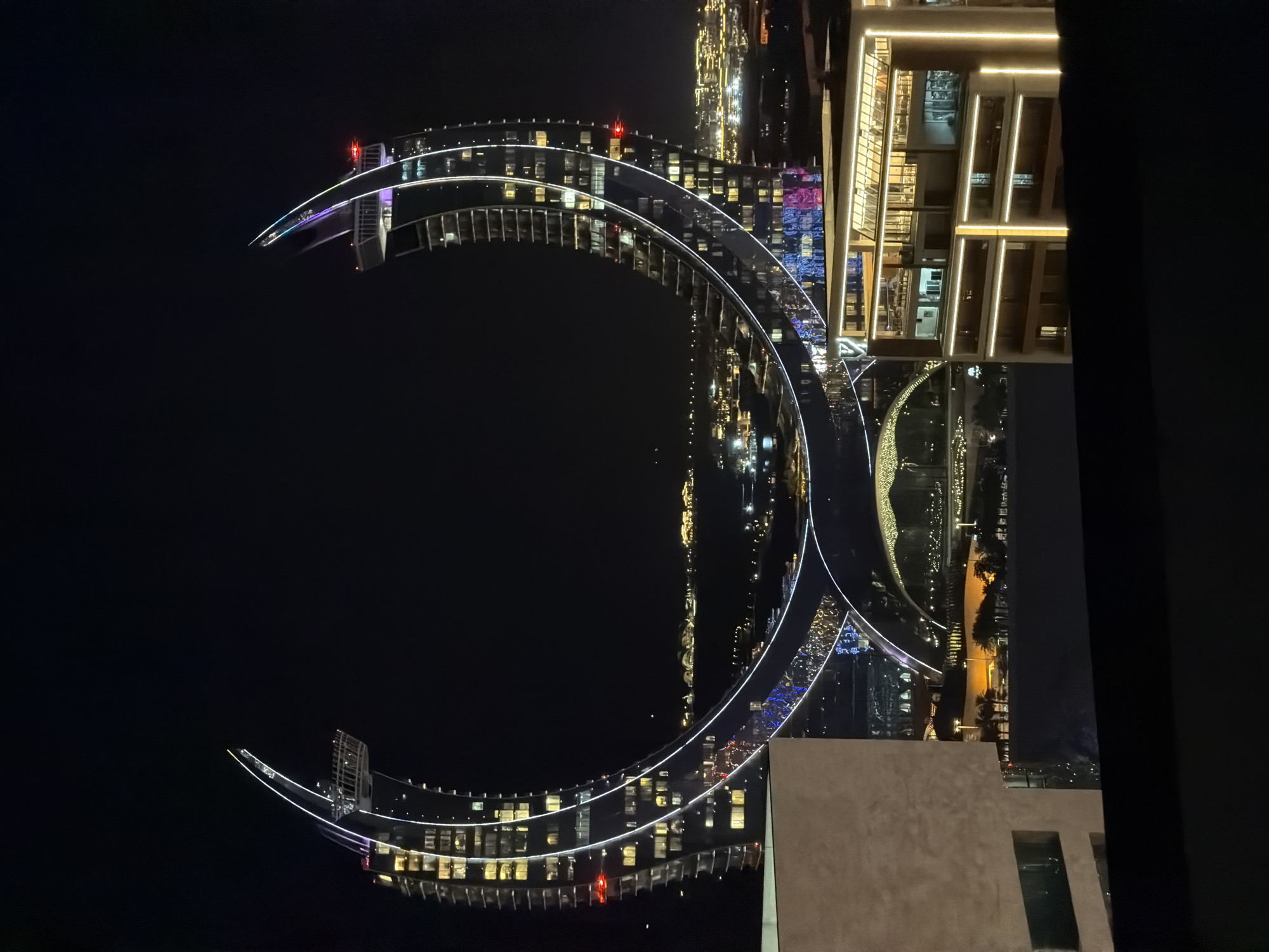 A nighttime aerial view captures the striking architecture of the Katara Amphitheatre and surrounding buildings in Doha, Qatar. The focal point is a colossal, crescent-shaped building illuminated by a cascade of white and colored lights that trace its elegant curves. This distinctive structure features multiple levels and a central open space, suggesting a unique design that embraces its urban surroundings.
Below the crescent building, the Katara Amphitheatre stands as a prominent landmark. Its curved seating arrangement is visible, and the structure itself appears to be a blend of modern and traditional architectural styles. Lights accentuating its form suggest it is illuminated for an event or as a general nighttime display.
In the foreground, a contemporary building with clean lines and bright, warm lighting provides a contrast to the more dramatic forms in the background. This building has multiple floors with visible interior spaces and external lighting that outlines its geometric structure.
The background reveals a sprawling cityscape dotted with numerous buildings and lights, indicating a densely populated urban area. The lights of distant structures suggest a vibrant city at night, stretching towards a dark horizon. The overall scene is characterized by sophisticated architectural designs and extensive illumination, highlighting the modernity and visual appeal of Doha's urban landscape.