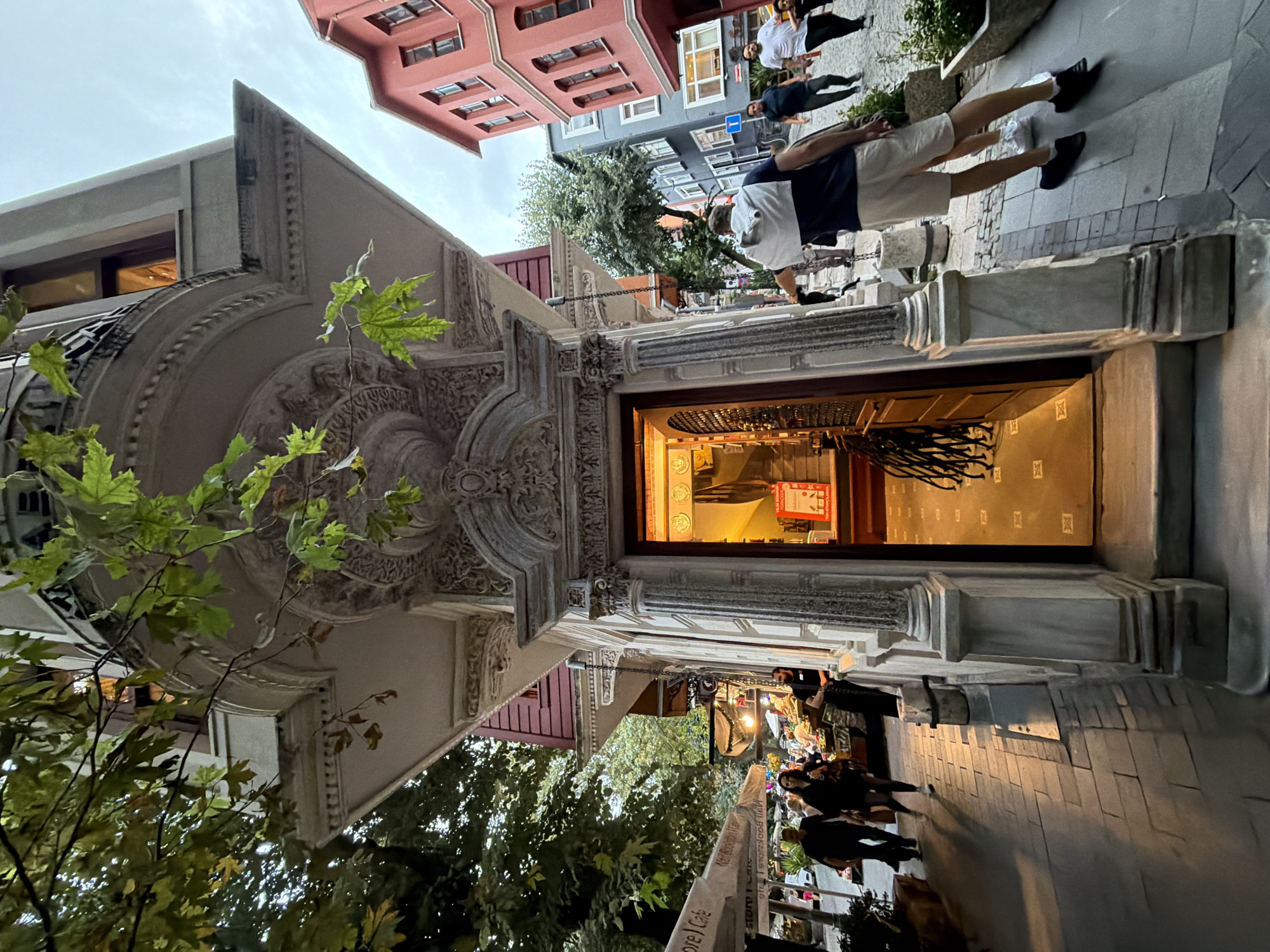 The image depicts a street scene in the Üsküdar district of Istanbul, Turkey, at dusk. A building with ornate white stone detailing and a curved balcony is prominent in the foreground, with an open doorway revealing a warmly lit interior. To the left, trees with green and yellowing leaves overhang a cobblestone sidewalk lined with businesses, one of which has umbrellas and outdoor seating. Several people are walking on the sidewalk, some carrying shopping bags. To the right, a red building with multiple windows stands beside a grey building. A man in a white and navy shirt and light shorts is standing with his back to the camera, looking towards the street. Other individuals are visible further down the street. The overall atmosphere suggests a lively urban environment transitioning into evening.