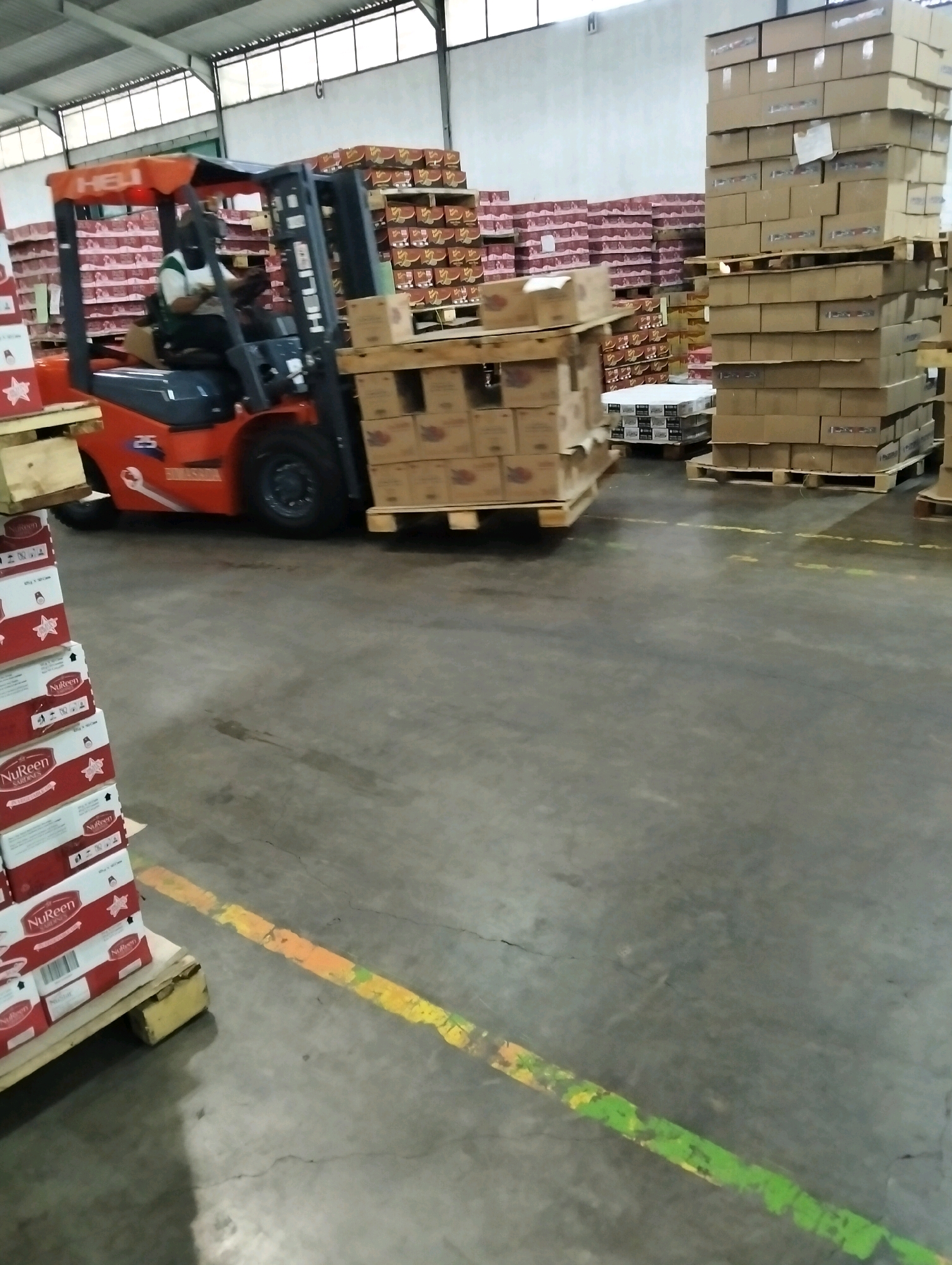 In a warehouse located in Tanggulangin, Indonesia, a forklift operator wearing a green and white shirt and a dark mask is driving an orange forklift. The forklift is carrying a pallet stacked with cardboard boxes. The warehouse is filled with numerous tall stacks of cardboard boxes, some of which are labeled with red and white branding. The floor is a concrete surface with painted yellow and green lines.