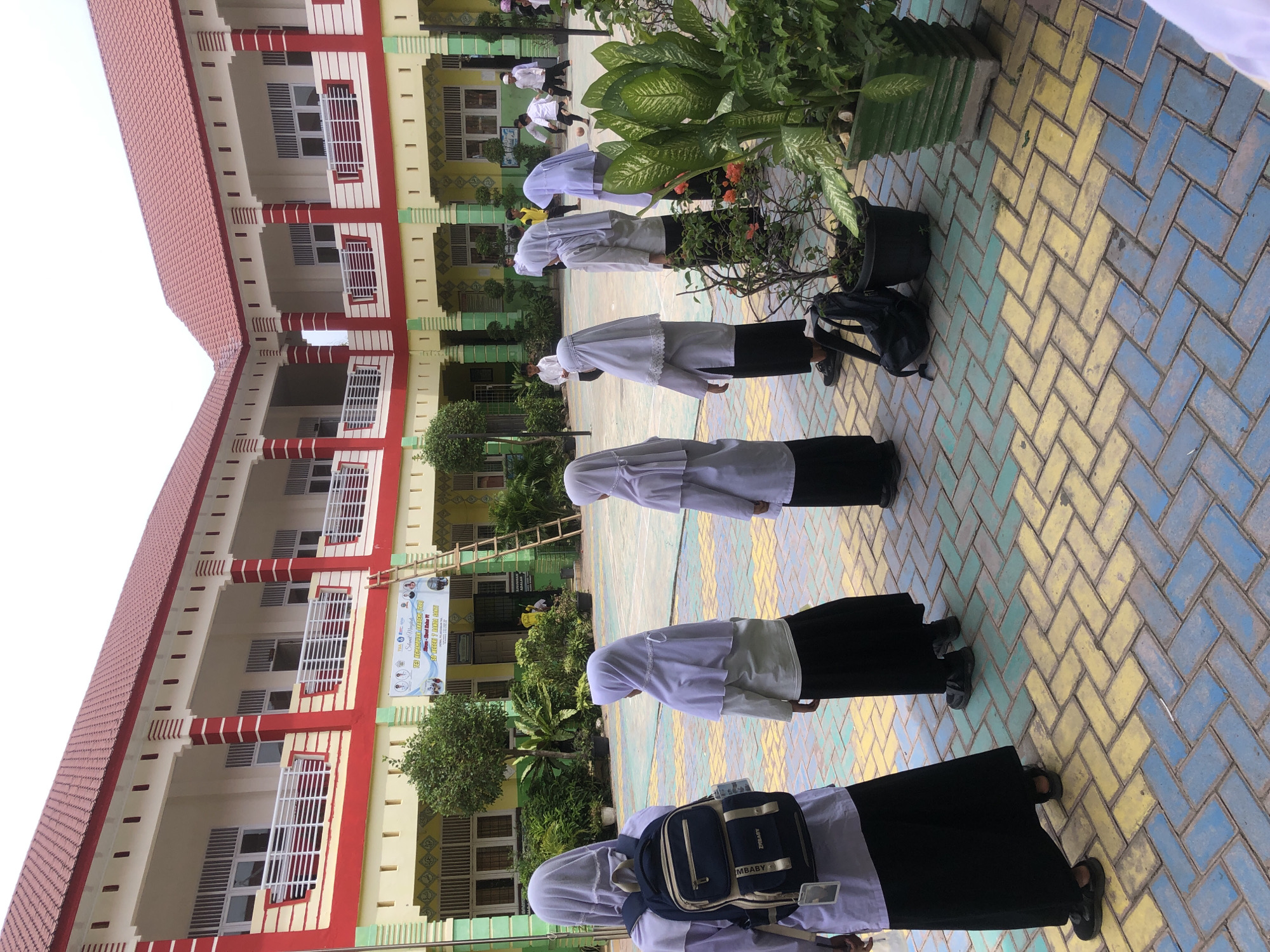 The image depicts an outdoor courtyard at a school in Lhokseumawe, Indonesia. Several young people, most wearing white hijabs and dark skirts or pants, are visible. Some are standing in lines, while others are engaged in activities in the background, including what appears to be a game of soccer. The courtyard is paved with colorful bricks in patterns of yellow, blue, and green. A two-story building with red roofing and white and red trim surrounds the courtyard. A ladder leans against the building, and a banner with text is displayed on the facade. Plants in pots are arranged around the area. The scene suggests a school day, possibly during recess or a break.