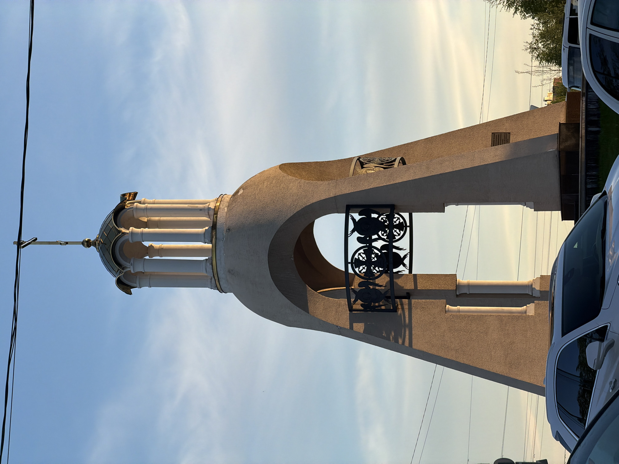 The image shows a tall, arched structure, likely part of a religious building or monument, possibly a church bell tower. The structure is light-colored and features a domed top with a cross. There are decorative pillars and a metal grill with symbolic designs in the archway. The structure appears to be located in an urban or suburban area, with cars parked nearby and power lines visible in the background. Based on the location specified, it's assumed that this structure is in Tokkari, Russia.