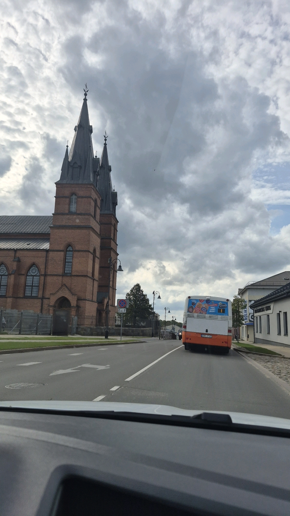 An exterior, eye-level, wide shot captures a brick church with tall spires on the left of the frame. The church has arched windows and a dark, pointed roof. A bus with an advertisement on its back is driving away from the camera down a road to the right of the church. The bus is white with an orange lower half. A license plate on the bus reads "KM 3169". The road has lane markings and is in front of a sidewalk and buildings. The sky is cloudy. This image appears to be taken in the city of Rezekne, Latvia.