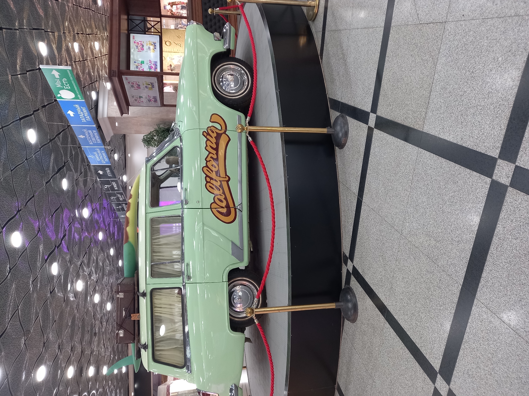 A pale green vintage station wagon, likely a GAZ-21 Volga, is displayed on a black circular platform within a commercial establishment. The car has a surfboard and luggage strapped to its roof rack. The word "California" is emblazoned in a stylized font on the rear passenger door. Red rope barriers, supported by gold stanchions, surround the platform. The background features a patterned ceiling with circular lights and directional signage in Russian, indicating parking and a cafe. The floor is tiled in a grey and black checkered pattern. The scene is likely in Yekaterinburg, Russia, given the Russian signage. No people are visible in the immediate vicinity of the car.
