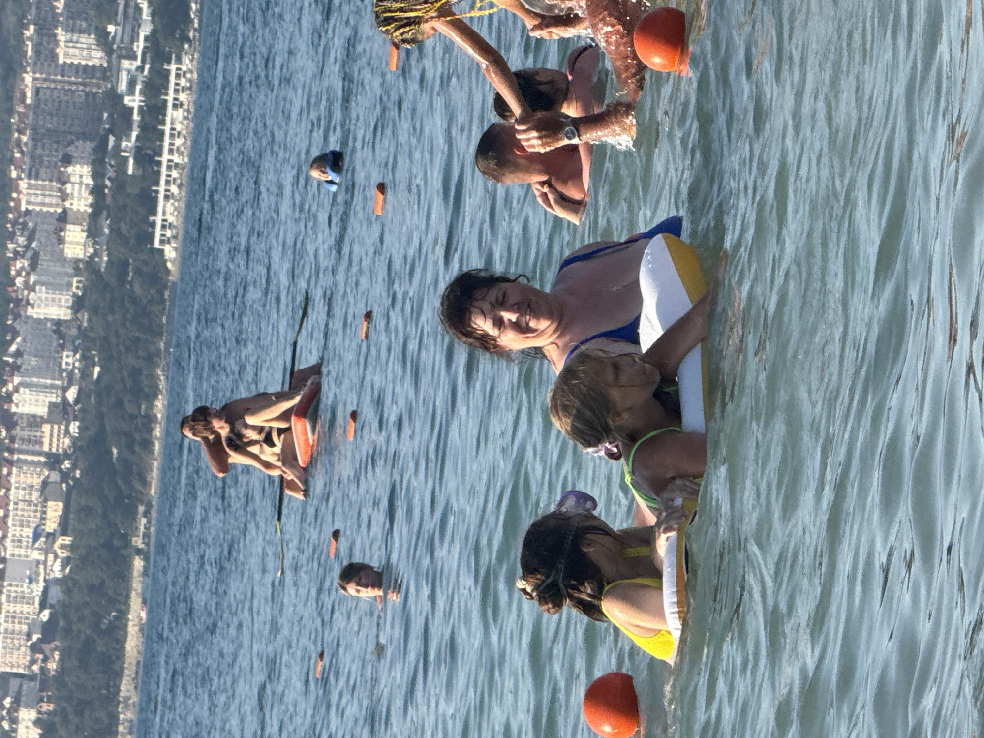 The image depicts a group of people enjoying a sunny day at the beach in Gelendzhik, Russia. In the foreground, a woman with dark hair and a blue bathing suit is smiling, floating in the water with two children. One child, wearing a yellow swimsuit, is on an inflatable raft. Another child, wearing a green swimsuit, is also on an inflatable raft with goggles on their head. In the middle ground, a couple is embracing on a floating device, and a child in a life vest is visible further out in the water. In the background, the coastline of Gelendzhik is visible, with buildings and lush green hills. The sea is calm and blue, with small waves. Orange buoys are scattered throughout the water, marking a swimming area.