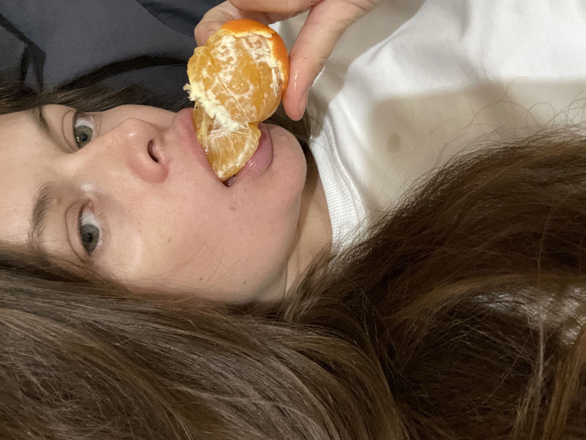 The image shows a close-up of a woman's face as she is about to eat a peeled tangerine. She has long, dark brown hair that is partially obscuring her face. Her right eye is visible, with a green iris. Her lips are slightly parted as she brings the tangerine to her mouth. The tangerine is peeled and appears to be broken into two segments, with one segment already partially in her mouth. Her fingers are holding the tangerine. She is wearing a white shirt. The background is dark and out of focus, suggesting an indoor setting. There is no identifiable location in the city of Заречный, Russia, visible in this image.