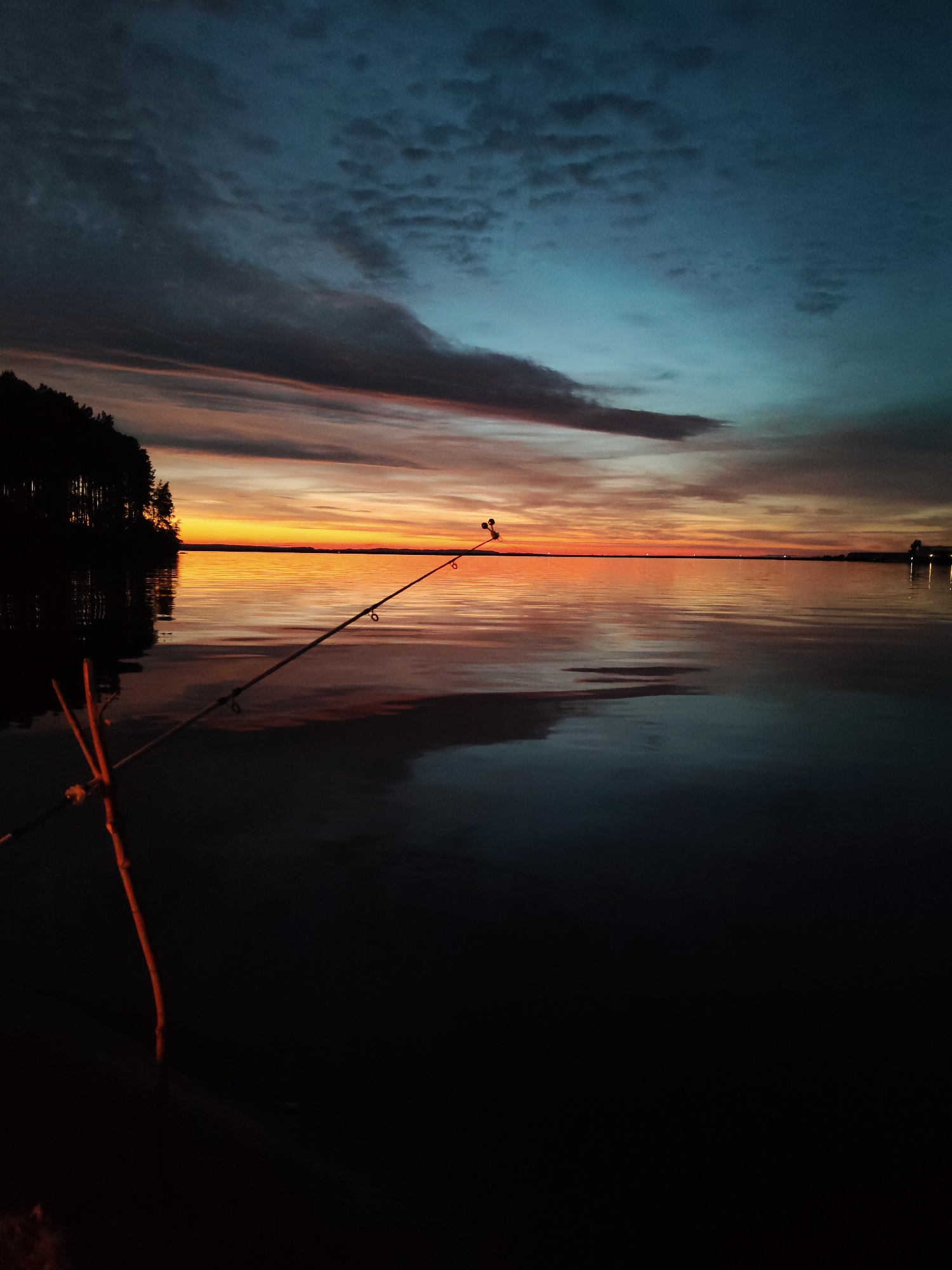 A fishing rod rests in a wooden holder on the edge of a body of water. The sun is setting, casting a warm glow across the water and sky. The sky is a gradient of blues and oranges, with clouds scattered throughout. A line of trees is visible on the left side of the image, silhouetted against the sunset. The water is calm and reflects the colors of the sky. In the distance, there are lights from what appears to be a town or buildings.