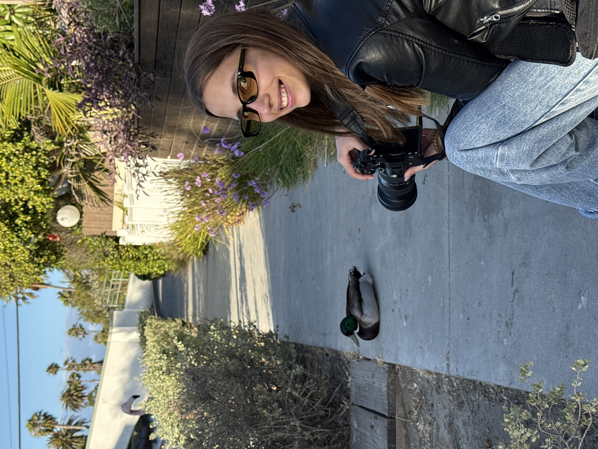 A person wearing a black leather jacket, light blue jeans, and glasses is holding a camera and smiling. The person is looking down at a duck that is on a sidewalk next to some greenery. The location appears to be in Los Angeles, United States of America. In the background, there are houses, palm trees, and a clear blue sky.