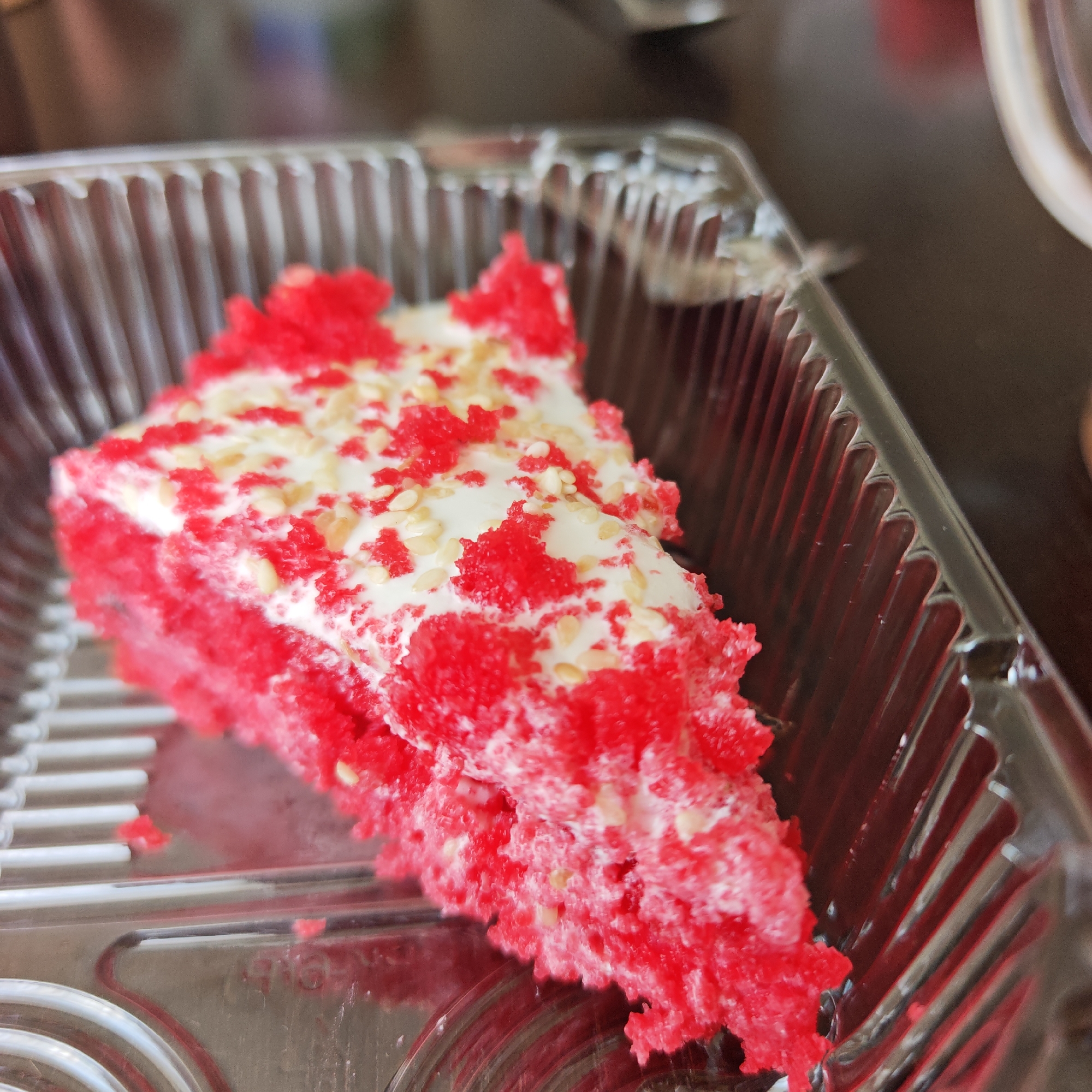 A slice of red cake with white frosting and scattered seeds rests in a clear plastic container. The container is ridged. The surface underneath is dark. The location and any people are not discernible from the image.