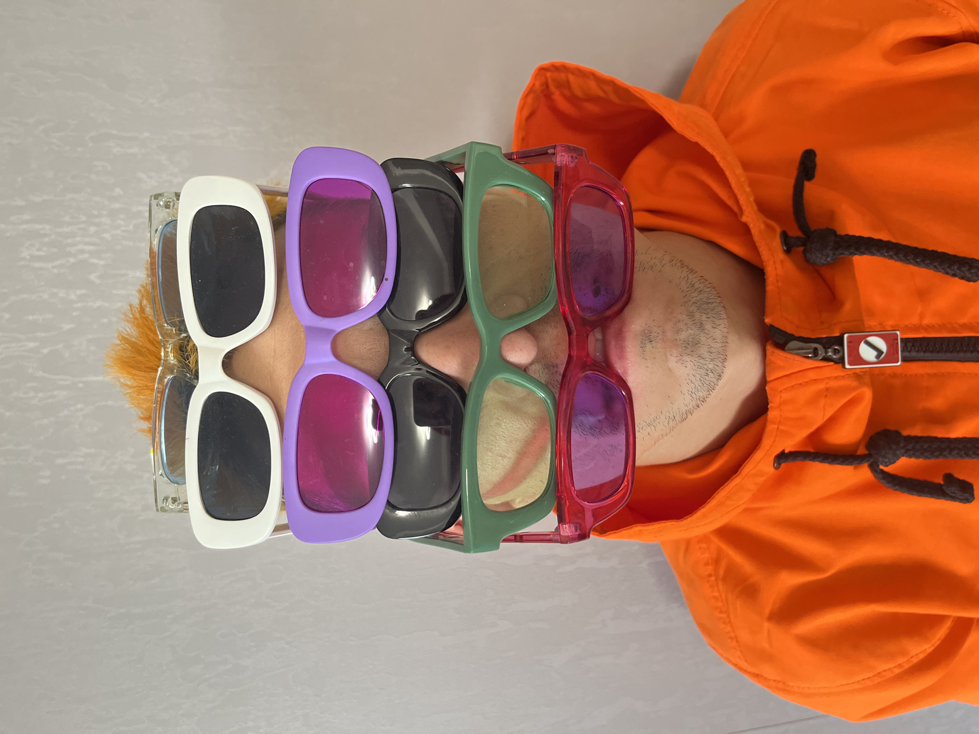 The media file features a close-up shot of a person wearing multiple pairs of sunglasses stacked on their face. The subjects are a person, whose face is partially obscured by the sunglasses, and six pairs of sunglasses with varying frame colors: clear, white, purple, black, green, and red. The person is wearing an orange hooded jacket with drawstrings. The scene is likely indoors, given the lighting and the plain background. The activity taking place is the person posing with the stacked sunglasses. Notable details include the different colors and styles of the sunglasses and the bright orange of the jacket. There is no visible text in the image. The location context provided is Chaikovsky, Russia.