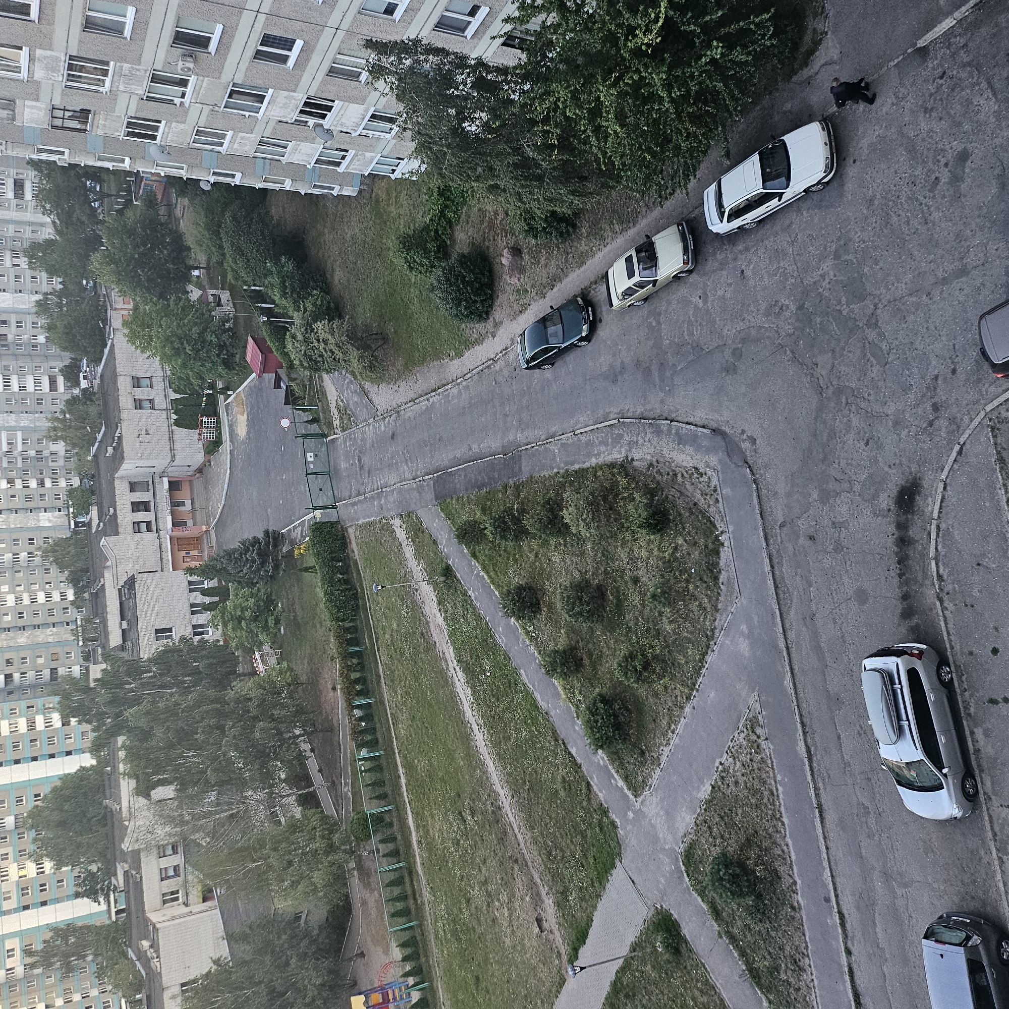 The image shows an aerial view of a residential courtyard in Brest, Belarus. The scene is dominated by apartment buildings, one of which is a tall, grey, multi-story structure on the right side of the frame with numerous windows. To the left and in the background, several other similar apartment buildings are visible. In the foreground, a grey SUV with a roof box is parked on the asphalt. Another grey SUV is parked next to it on the left. Further up the road, a light-colored sedan is parked, followed by a white sedan. A dark green sedan is parked further up this road. A person in dark clothing is walking away from the white sedan.
The courtyard area features paved paths and roads, grassy areas with scattered trees and bushes, and a playground in the upper left corner. A chain-link fence runs along the edge of the grassy area. A metal gate is visible on a road leading into the courtyard from the background. The overall environment appears to be a typical urban residential area. The lighting suggests it is either early morning or late evening.