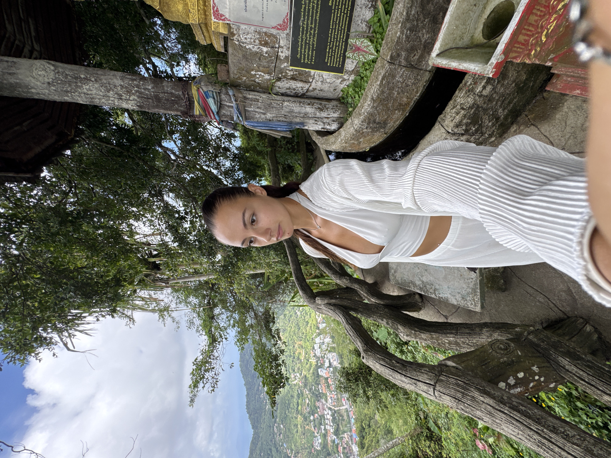 A woman in white stands on a hill overlooking Karon, Thailand. The woman is standing near a decorative wooden fence. Behind her, there are signs and Buddhist structures. The hillside is lush and green.