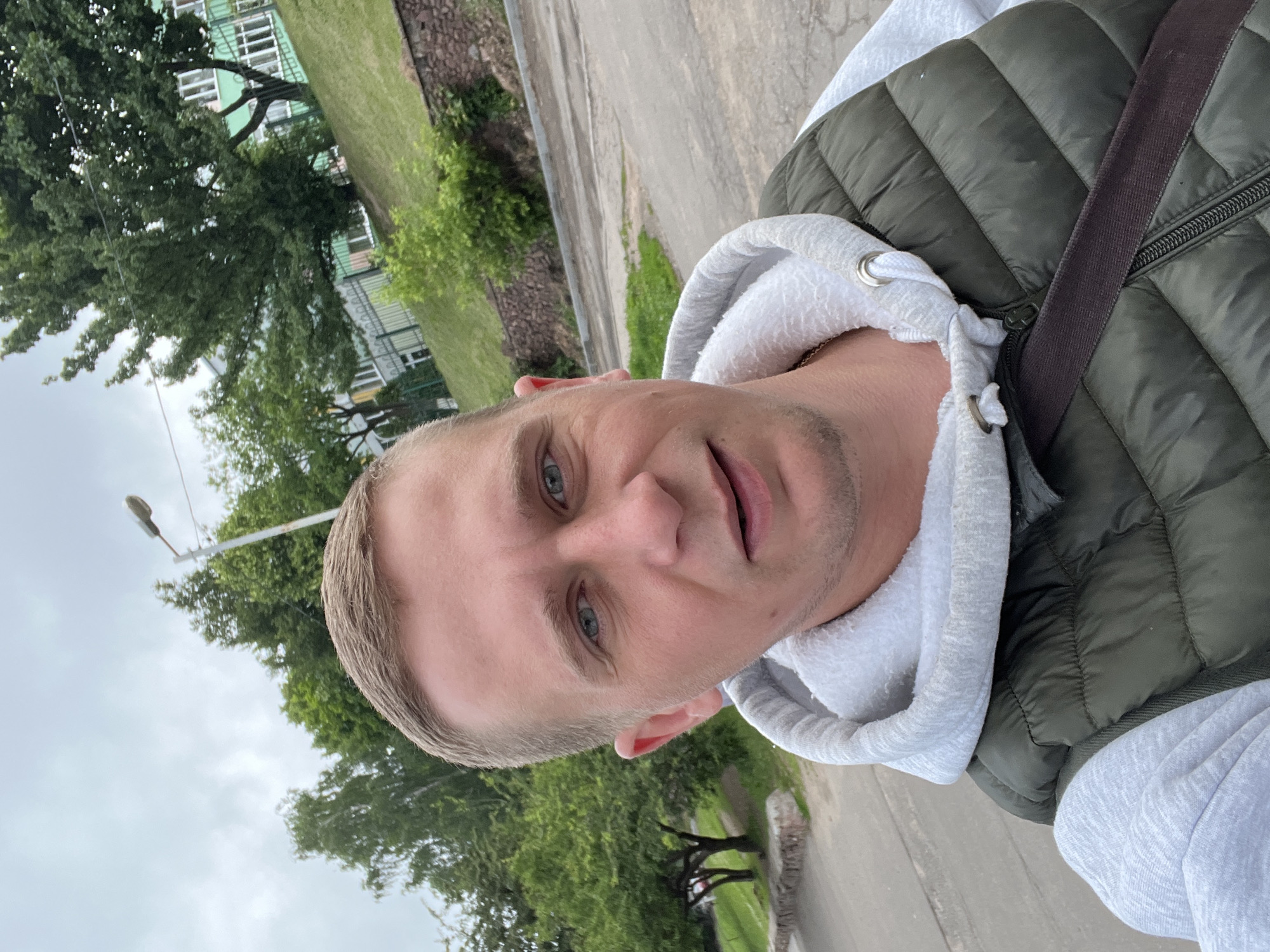 A close-up, outdoor selfie shows a fair-skinned man with short brown hair and blue eyes, wearing a light gray hoodie and a dark green puffer vest. He is looking at the camera with a neutral expression. Behind him, a tree-lined street is visible, with a grassy embankment and a building in the distance. The sky is overcast. The scene appears to be in a residential area.