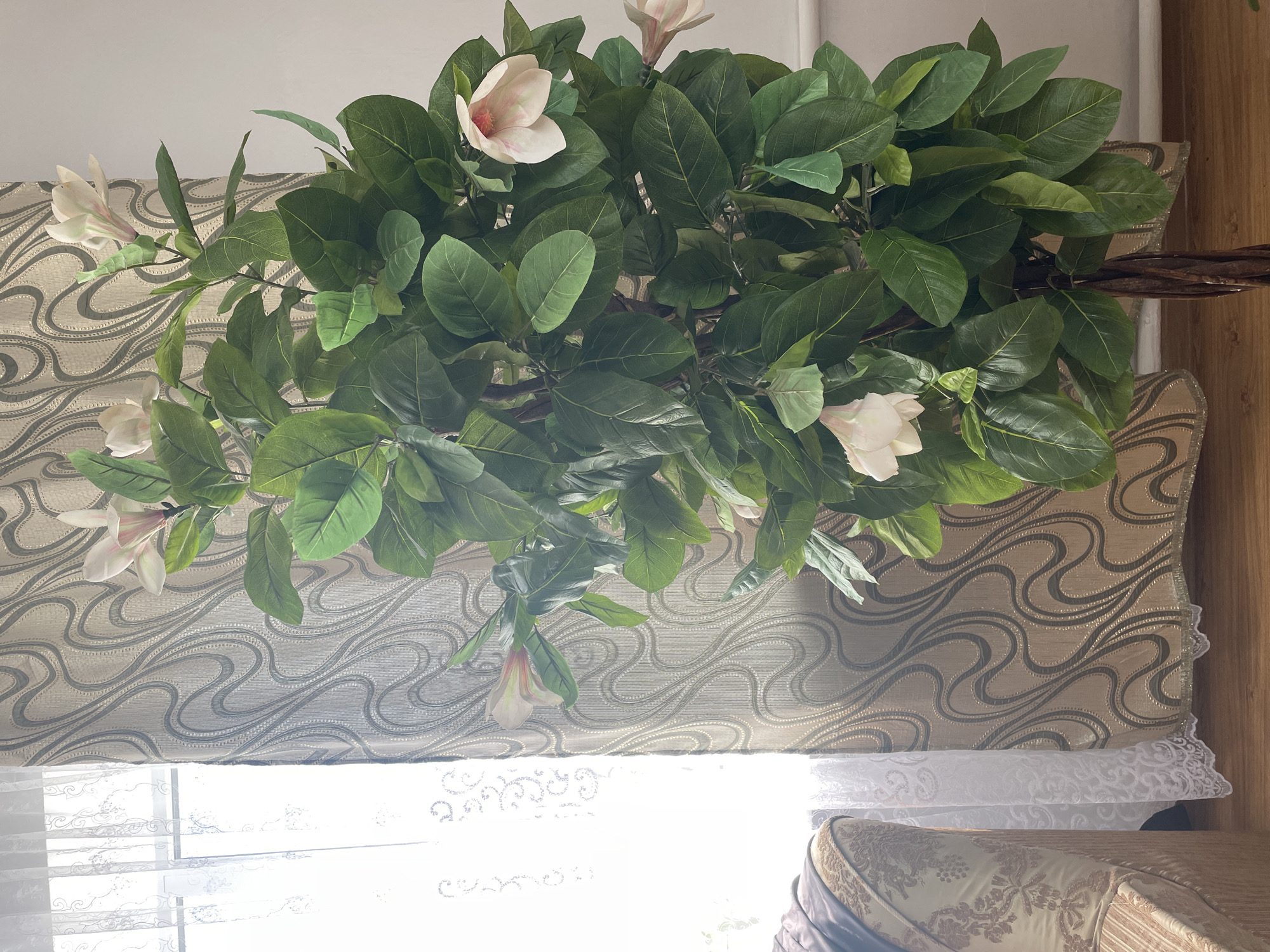 The image depicts an indoor scene, likely a living space, with a large artificial plant with green leaves and white and pink flowers as the central subject. The plant is situated in front of a patterned curtain with a swirling design in shades of beige and brown. To the left of the plant, a window is visible with sheer white lace curtains. A portion of an upholstered piece of furniture, possibly a sofa or armchair with a patterned fabric, is visible on the far left. The floor appears to be made of light-colored wood. The overall lighting suggests an indoor environment with natural light coming from the window. The location is identified as Shpykolosy, Ukraine. There are no people visible in the image.