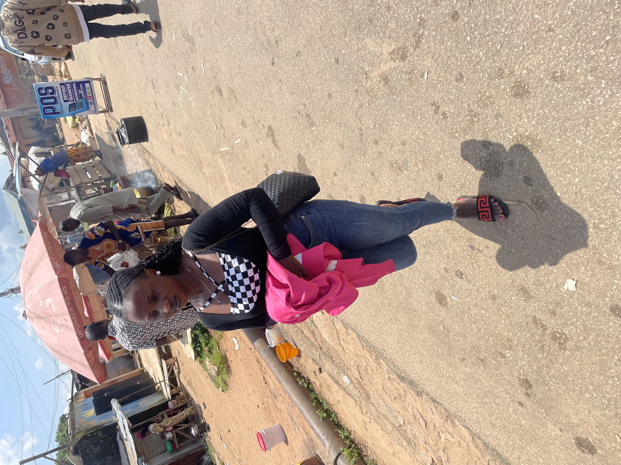 The image depicts an outdoor scene on a paved road in Jos, Nigeria. In the foreground, a young woman with braided hair is walking towards the camera, holding a bright pink garment. She is wearing a black long-sleeved top with a checkered pattern underneath, blue jeans, and sandals. A black quilted handbag is slung over her shoulder.
The background reveals a bustling street environment. Several people are visible, some standing and interacting, while others are engaged in various activities. Structures that appear to be market stalls or small shops line the side of the road, with some exhibiting corrugated metal roofing. An umbrella with the text "UPGRADE TO 4G" is prominently displayed. A blue sign with "POS SERVICES AVAILABLE" and details about deposit, transfer, and withdrawal is also visible. There are several buckets and other containers on the ground, suggesting a commercial or domestic setting. The overall atmosphere appears to be one of everyday street life in an urban area.