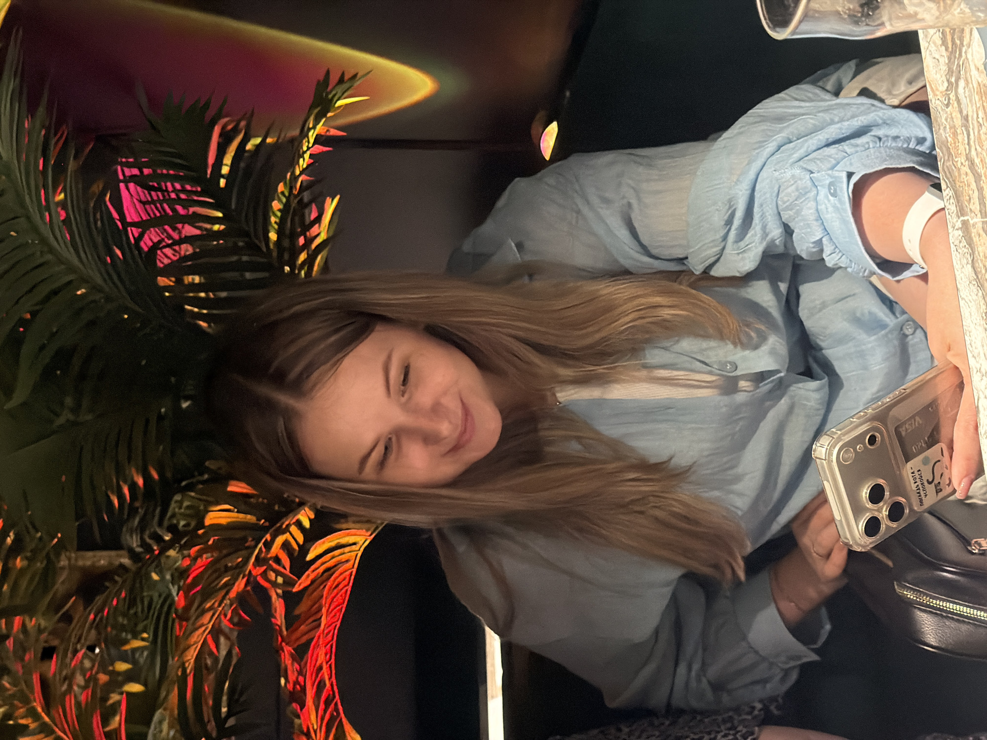 A young woman with blonde hair is seated at a table, looking down at her phone, which she holds in her right hand. She is wearing a light blue, long-sleeved shirt. Her left arm is resting on the table, and she has a white wristband on her left wrist. Next to her is a black purse with a gold zipper. Behind her, a large, leafy plant is illuminated by red and yellow lights, creating a vibrant backdrop. The scene appears to be indoors, possibly in a restaurant or bar, with a glass visible on the right side of the frame. The image was taken in the city of Chaikovsky, Russia.