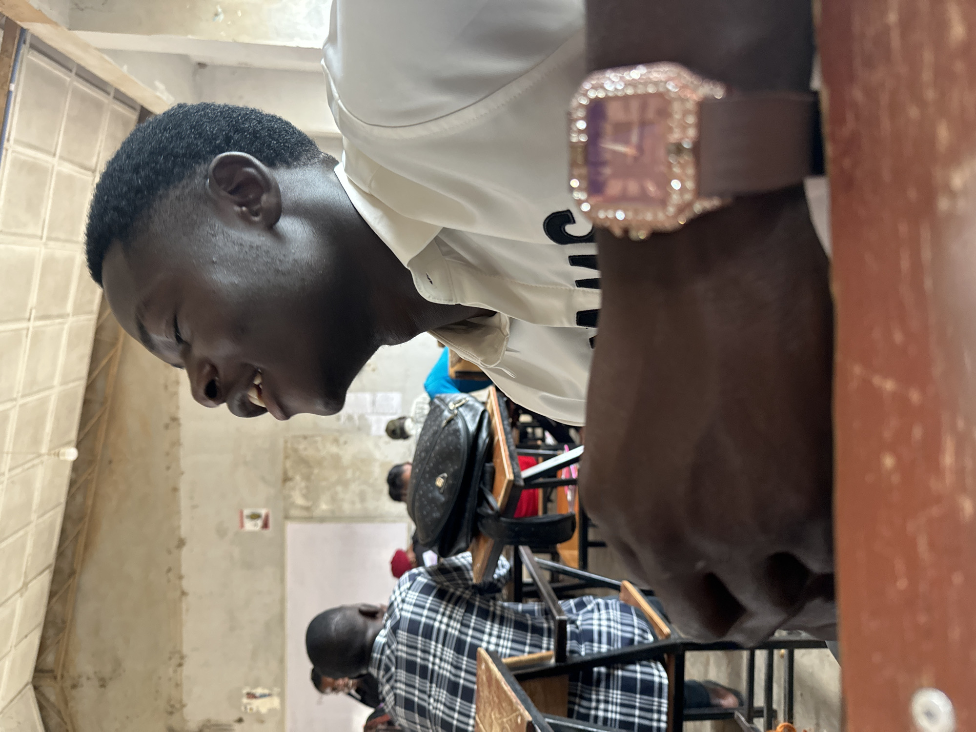 The image depicts a group of individuals inside a room with tiled walls, possibly a classroom, at an unspecified location in the city of Jos, Nigeria. A young Black man with a close haircut is visible in the foreground. He is wearing a white shirt. On his wrist is a square-faced watch with a brown band and what appears to be a jeweled or studded bezel.
In the background, several other individuals are sitting in chairs. One person is wearing a black and white plaid shirt. A black bag is hanging on the back of one of the chairs. The scene suggests a casual, possibly academic, setting.