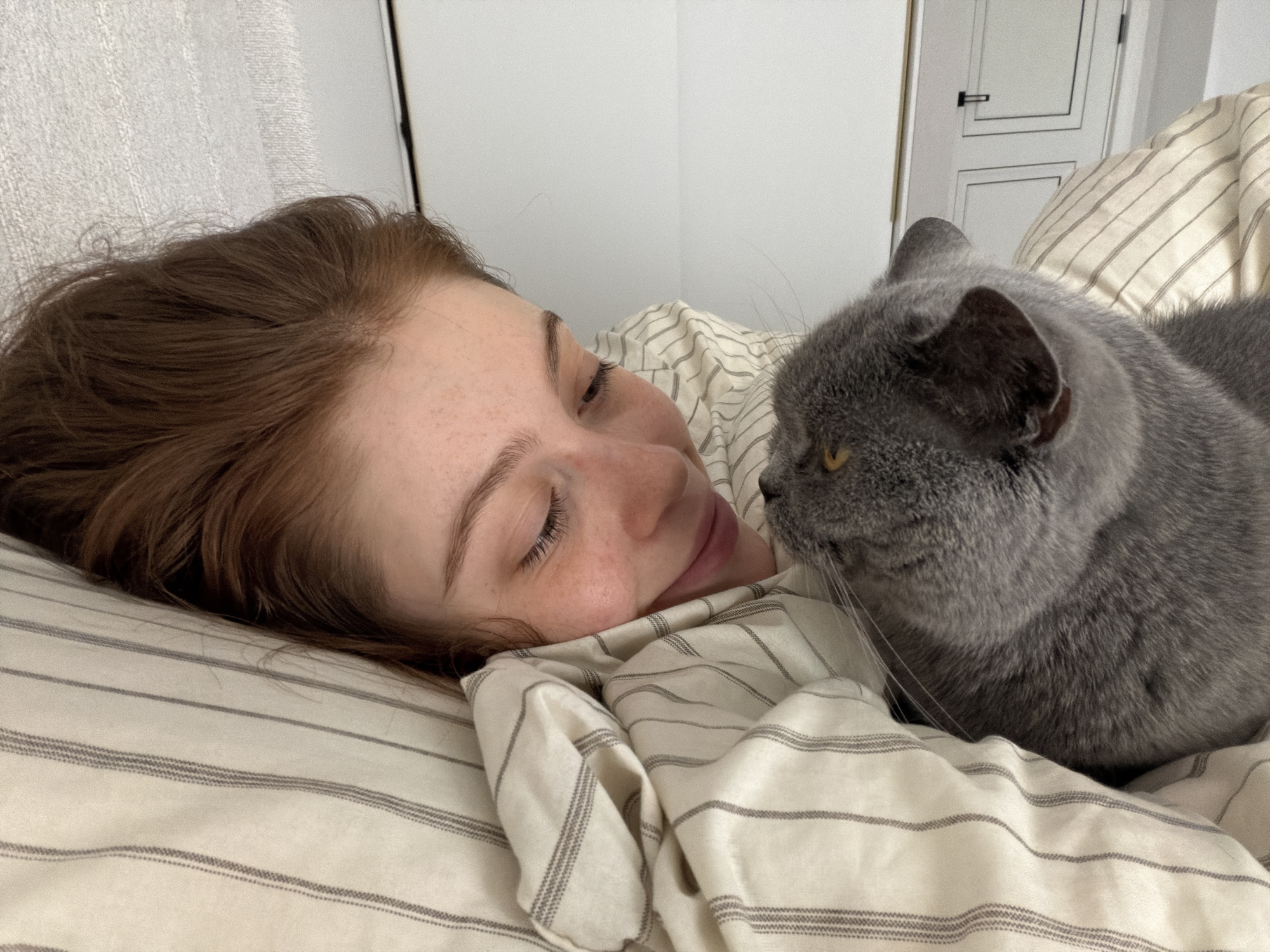 A woman with auburn hair lies on her side on a striped bed, facing a gray cat. The woman has freckles and closed eyes, and she is smiling. The cat is focused on the woman, close to her face. The background includes a white wall and a white door. The image does not provide enough information to determine the location in Tbilisi, Georgia.
