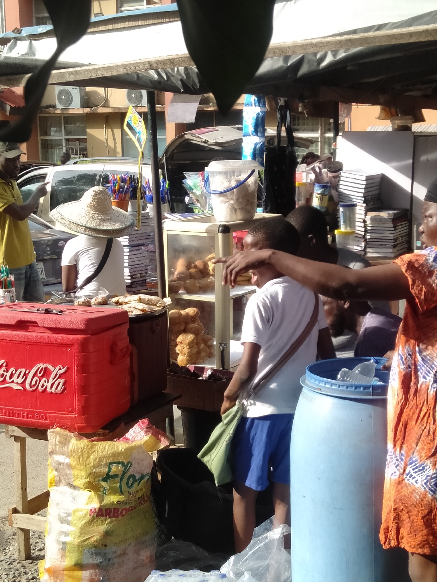 The image shows a street food stall in Lagos, Nigeria. A vendor wearing an orange patterned dress gestures with her right hand towards the stall, where a young boy in a white shirt and blue shorts stands with a green shoulder bag. The stall has a Coca-Cola branded cooler and a cabinet filled with fried dough. A man in a yellow shirt stands on the left, and another person wearing a wide-brimmed hat stands behind the stall, near pens and books. A "Flora" brand bag sits below the stall. A blue barrel stands near the vendor. There is a building in the background.